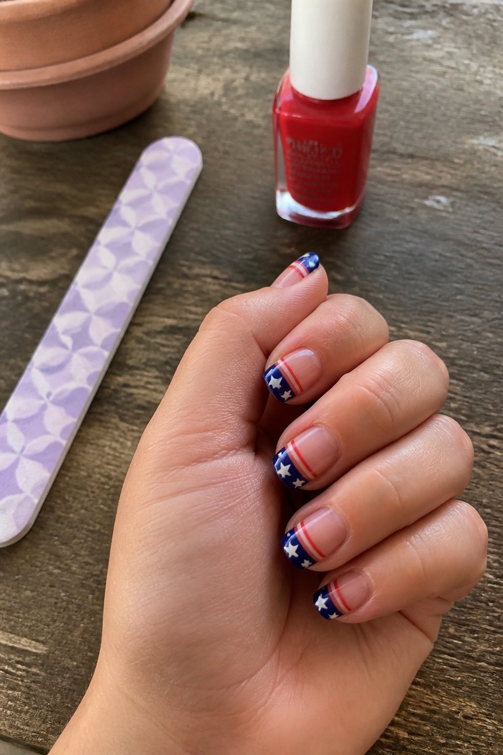 Close-up of a hand with simple 4th of July nails featuring a red stripe accent, resting on a wooden surface with nail care items nearby.