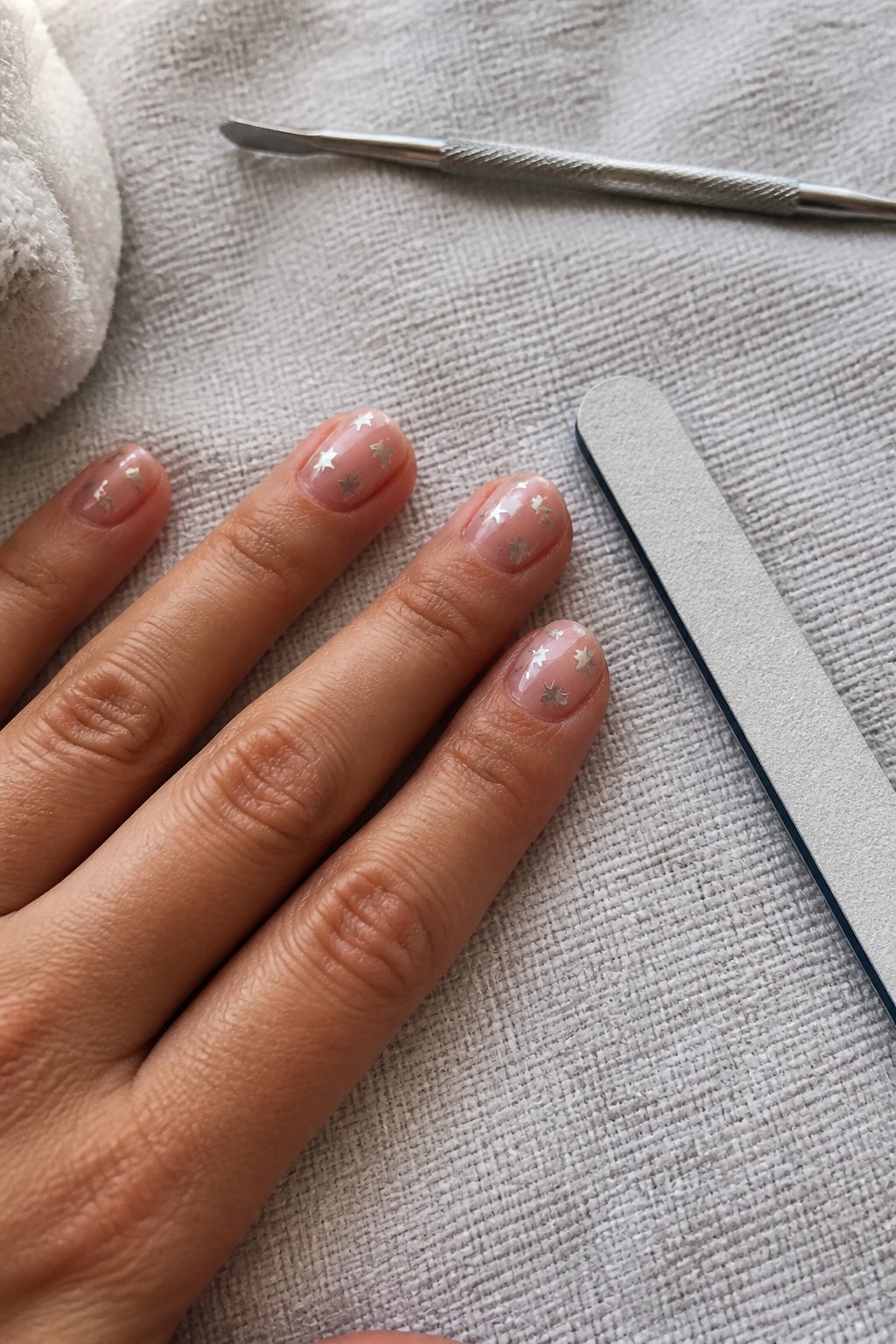 A close-up of a hand with simple 4th of July themed nails featuring silver star foil on sheer pink, resting on a textured surface with manicure tools nearby.