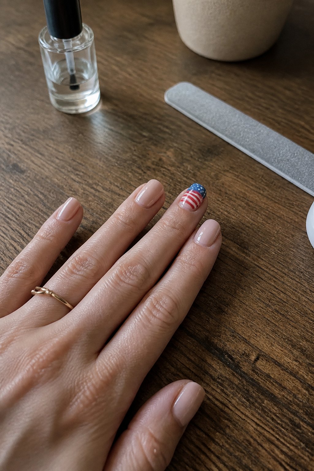 A hand with nude base nails and a small American flag accent on one nail resting on a wooden table with manicure tools nearby.
