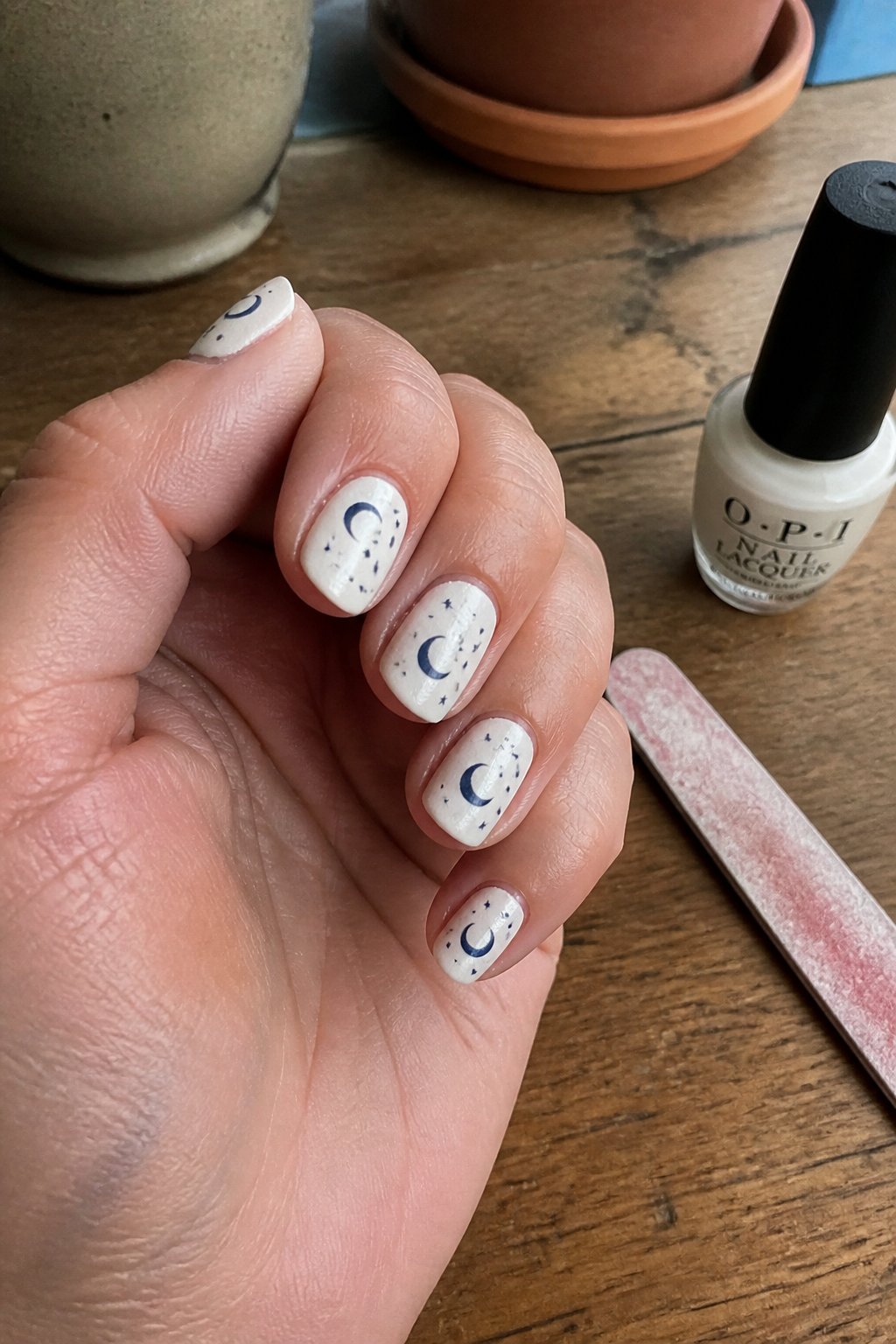 Close-up of hands showing pearl white nails with navy crescent moon designs, surrounded by nail care tools on a light surface.