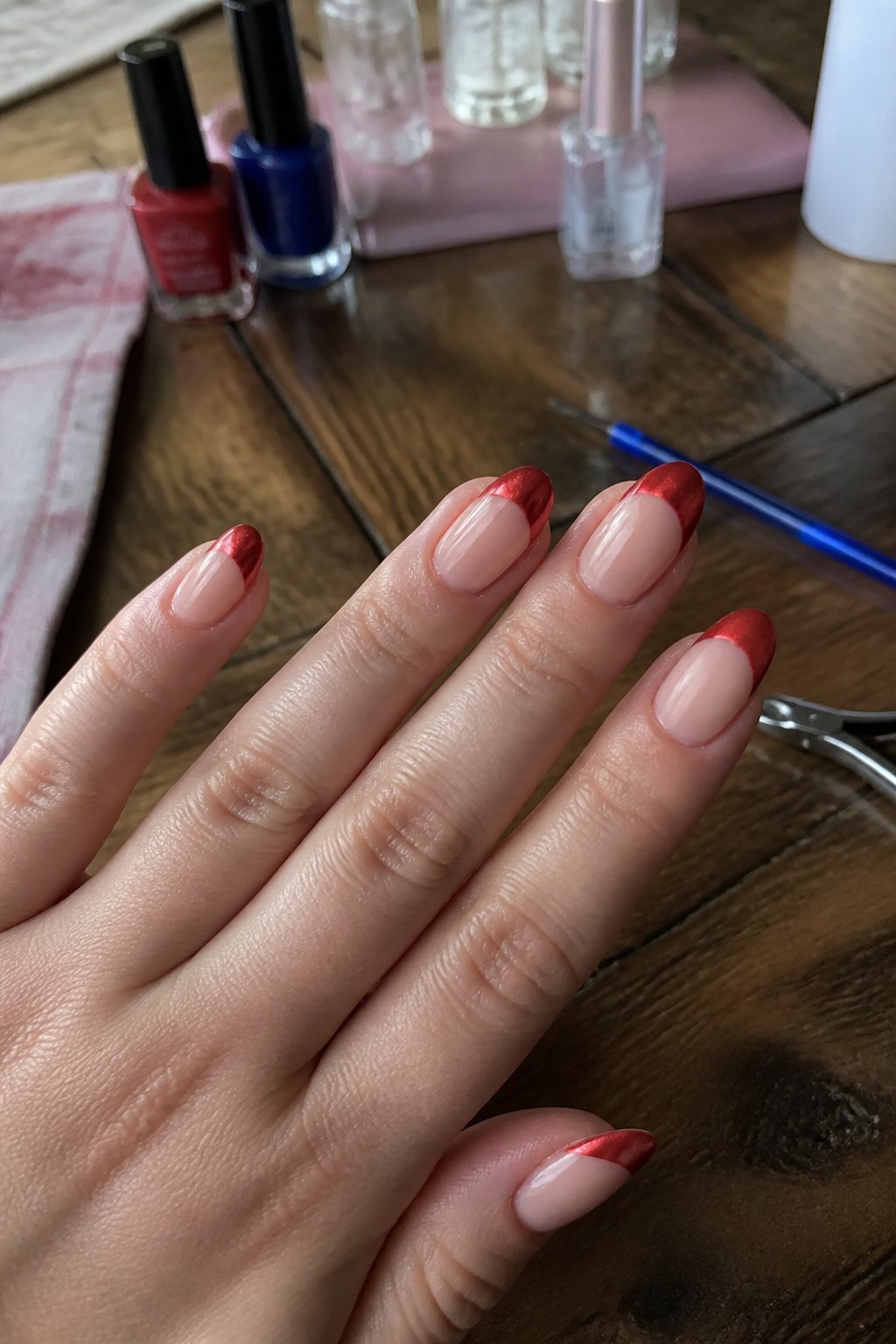 Close-up of natural almond-shaped nails with red chrome tips on a hand resting on a textured surface, surrounded by nail tools.