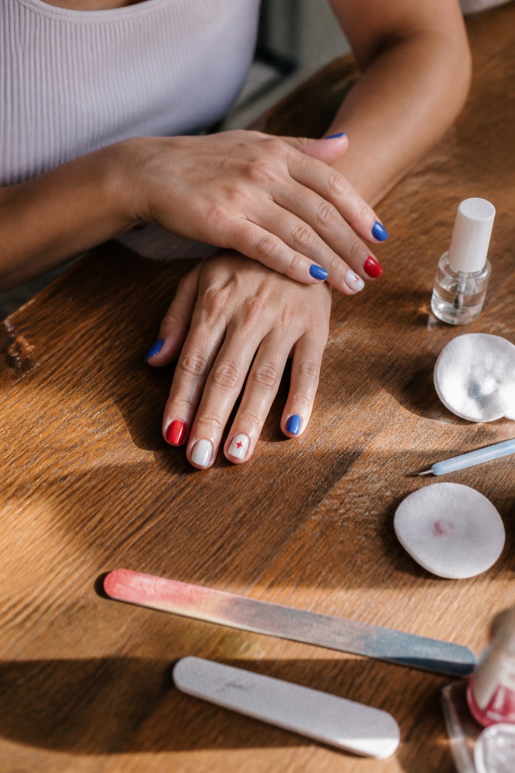 Close-up of hands with simple red, white, and blue nail designs on a wooden surface with manicure tools nearby.