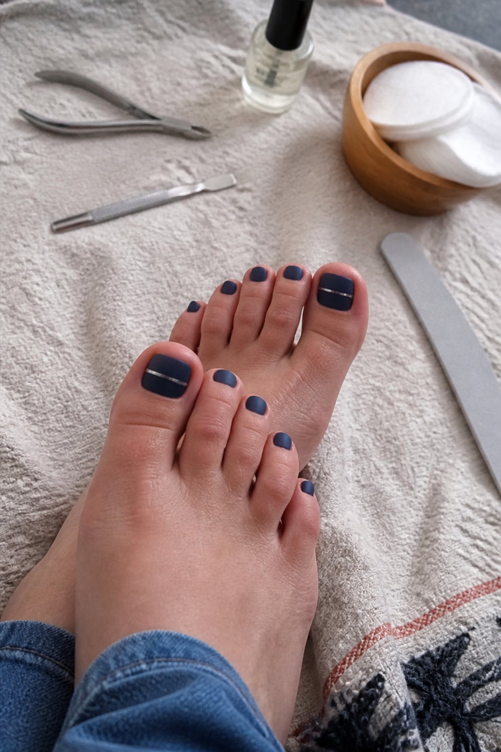 A close-up of feet with matte navy and metallic silver striped pedicure surrounded by pedicure tools on a light surface.