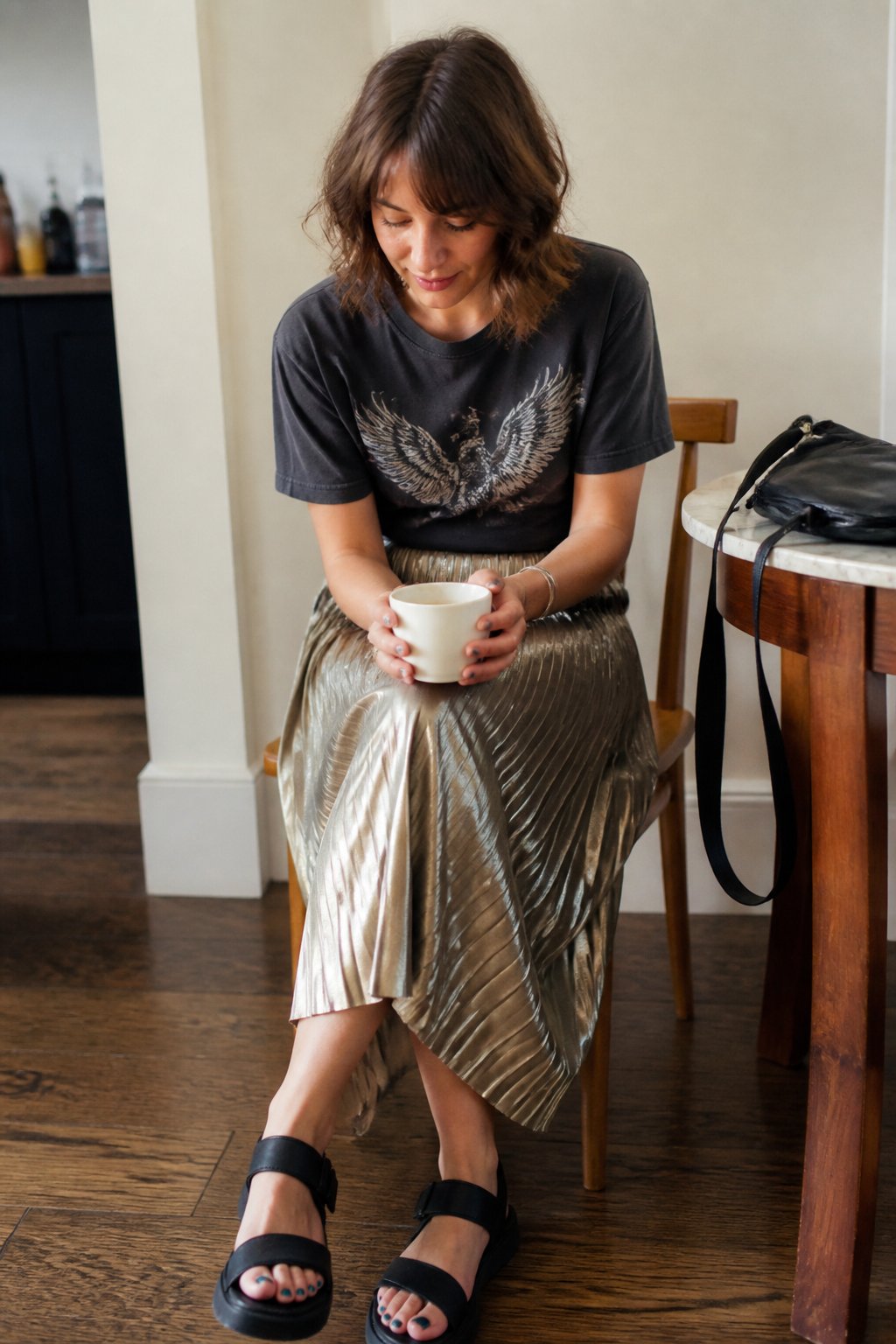 A woman wearing a metallic pleated midi skirt, graphic tee, and chunky sandals stands casually in soft daylight with everyday objects around her.