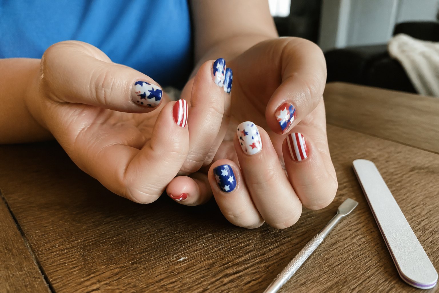 Close-up of hands with red, white, and blue Independence Day themed nails resting on a wooden table with manicure tools nearby.