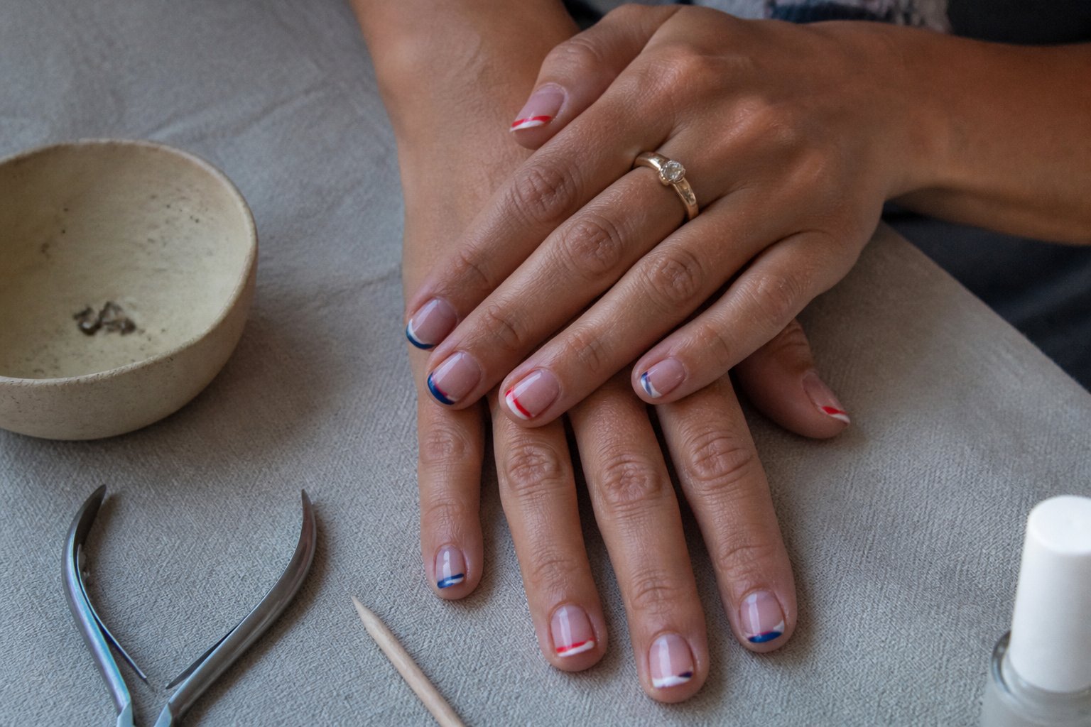 Close-up of hands with reverse French manicure featuring red, white, and blue tips, resting on a table with manicure tools nearby.