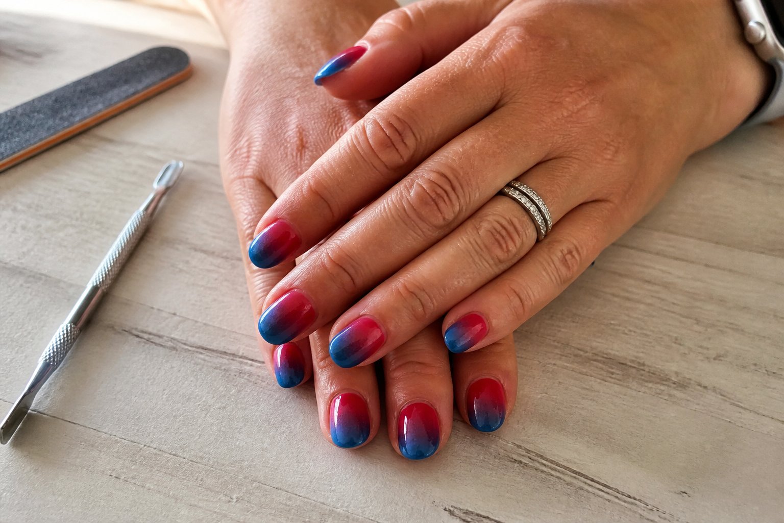Close-up of hands with red to blue gradient ombre nails resting on a wooden surface, surrounded by clean nail tools.