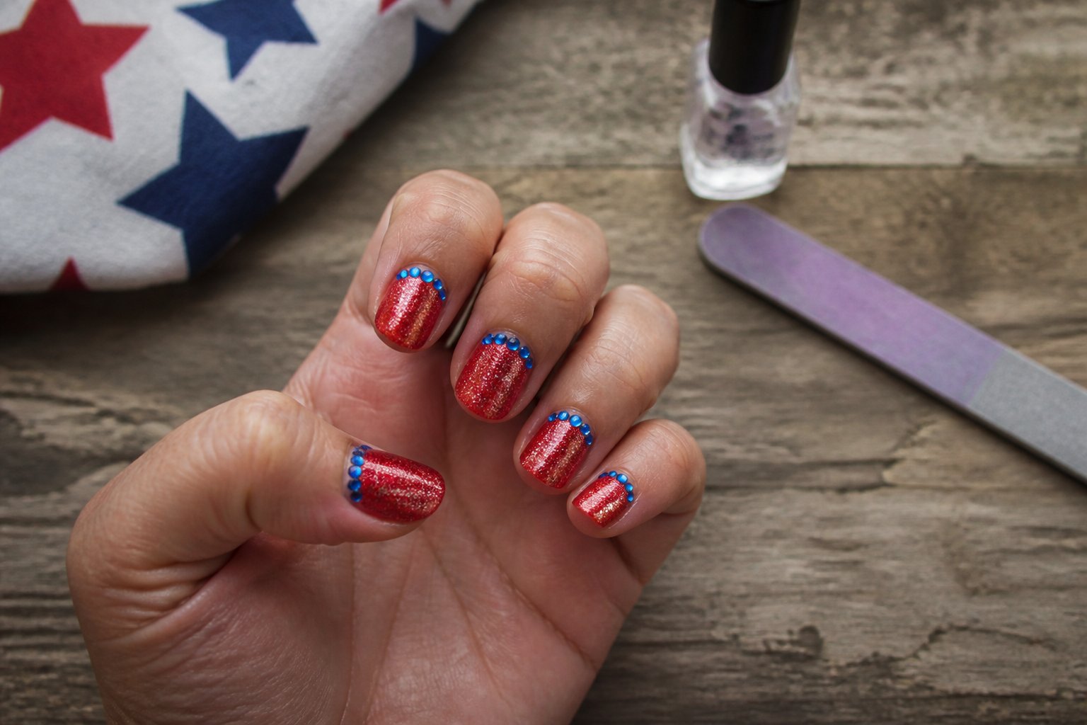 A hand with glittery red nails and a blue rhinestone crescent resting on a wooden surface surrounded by manicure tools.