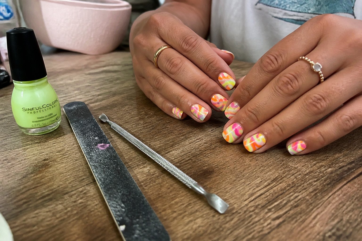 A close-up of hands with neon swirl nails holding nail care tools on a wooden table under soft daylight.