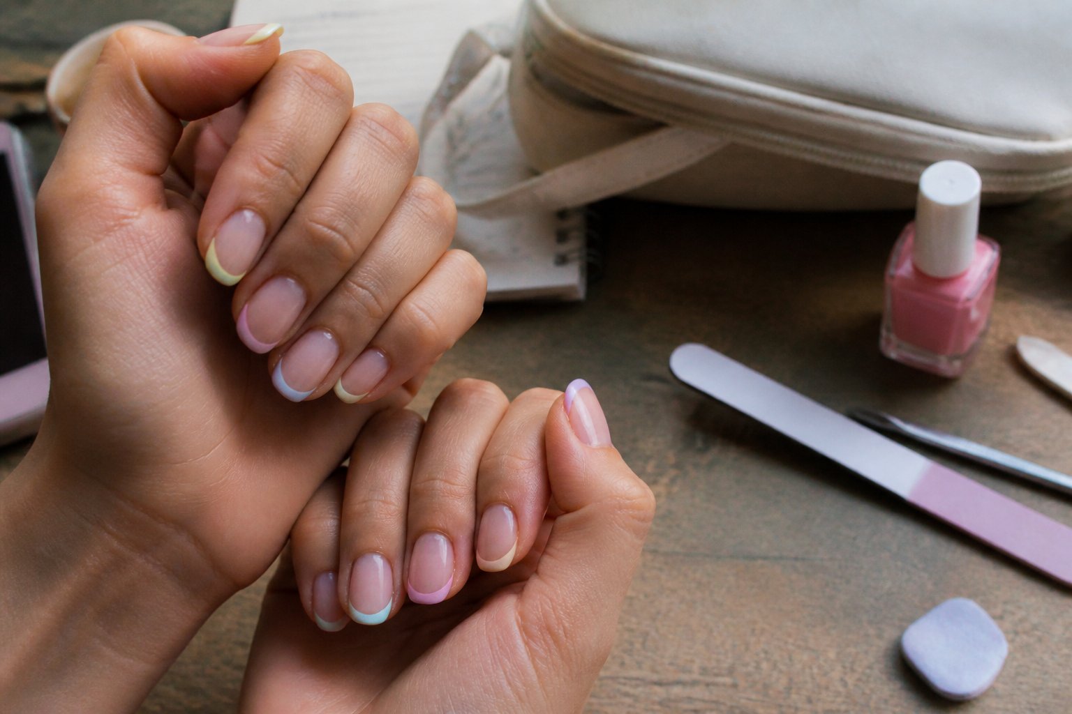 Hands with classic French manicure and pastel tips resting near manicure tools on a wooden surface in soft natural daylight.