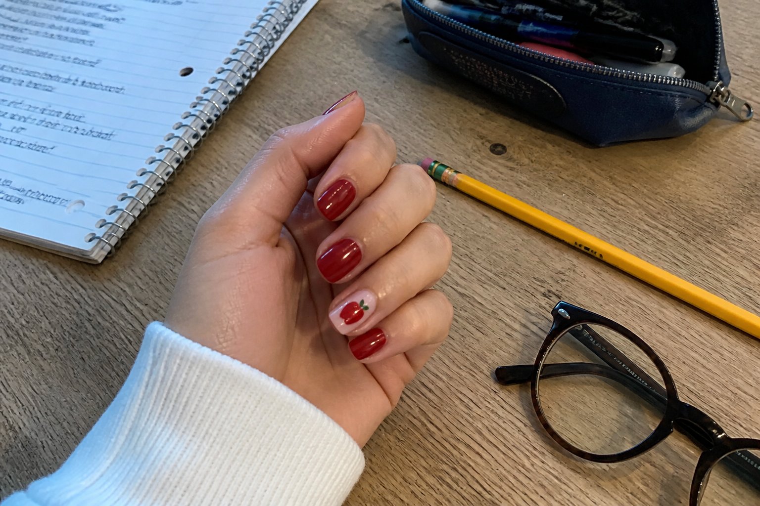 Close-up of a hand with glossy red nails featuring a tiny apple design, resting on a wooden desk with a notebook, pencil, and glasses nearby.