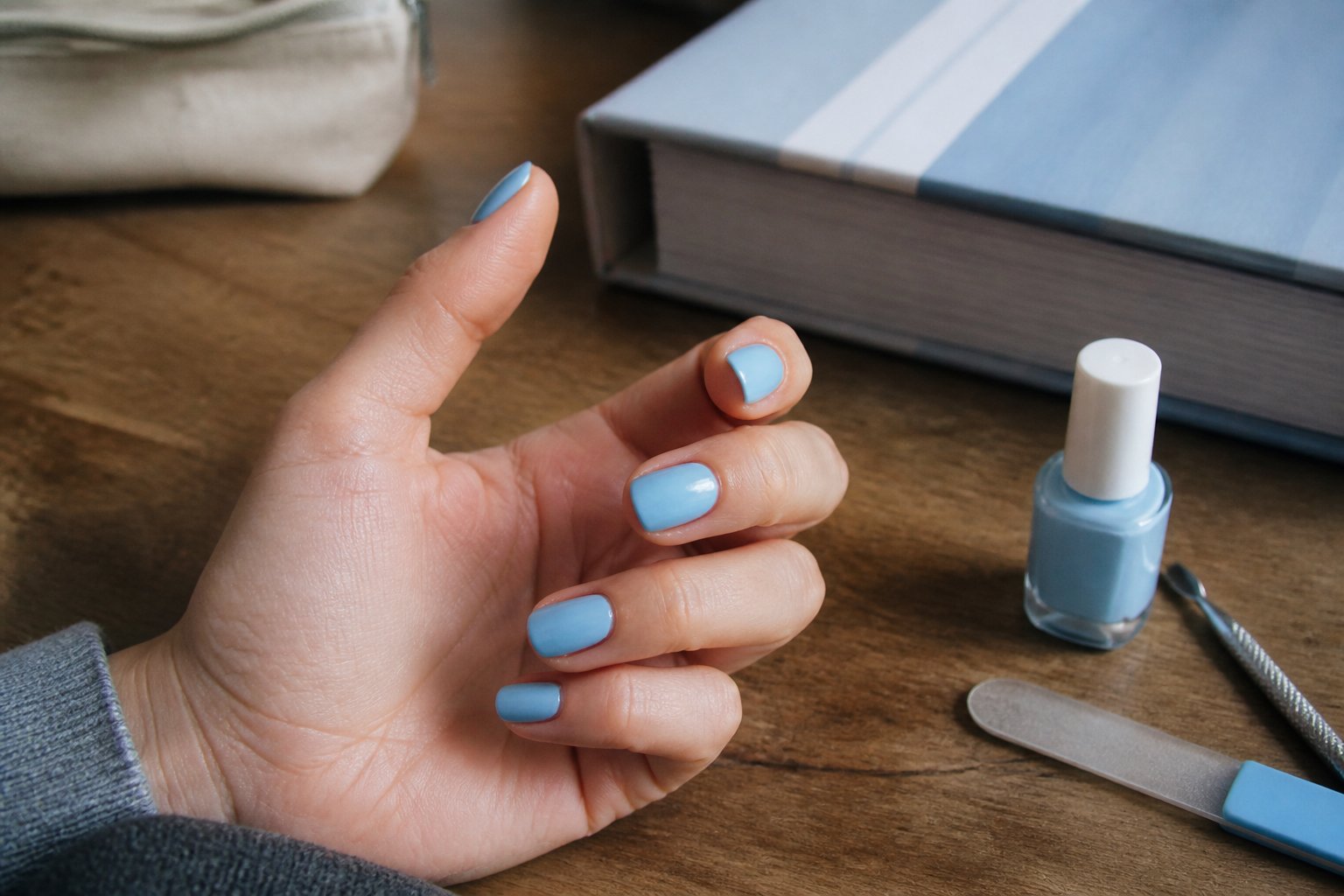 A hand with freshly painted nails near a pale blue textbook with a striped spine, surrounded by nail tools and a notebook on a table.