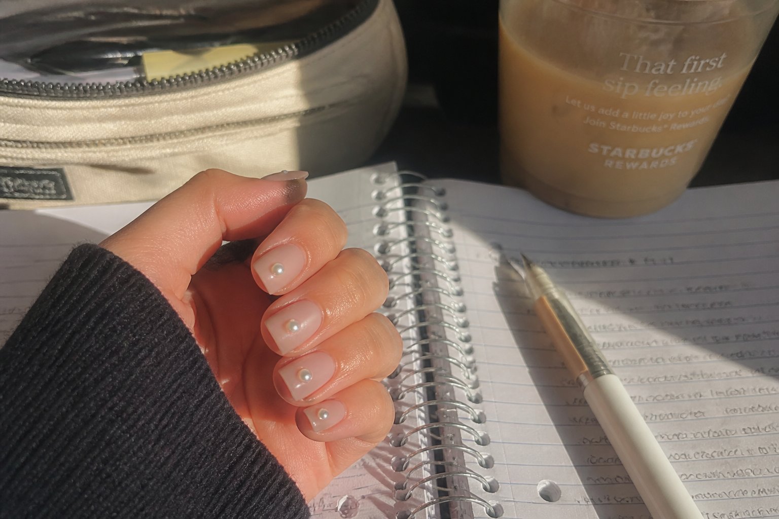 Close-up of hands with short square nails decorated with pearls, holding a slightly worn notebook and pen on a table with natural daylight.