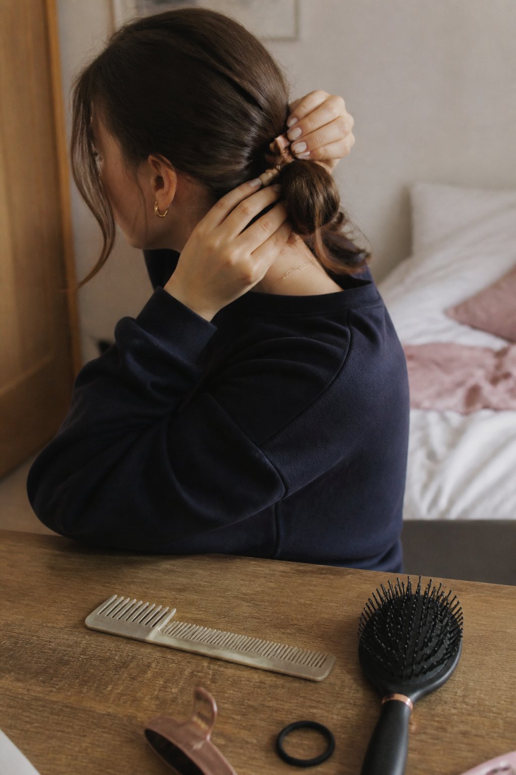 A person styling their hair into a messy low bun with face-framing pieces, with hands holding hair tools on a wooden table in soft daylight.
