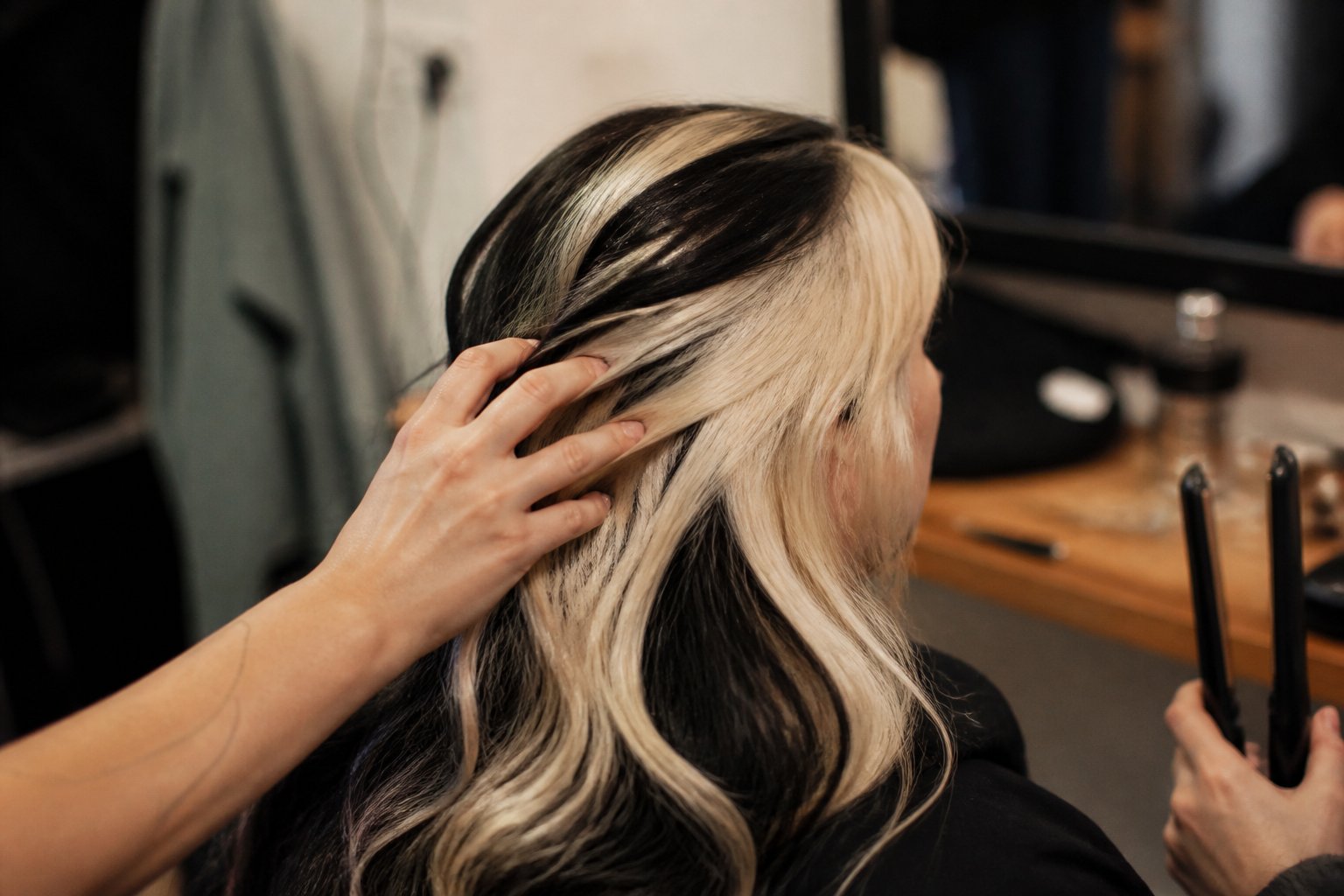 Close-up of a person with a black-and-platinum skunk stripe hairstyle, with hands and hair tools nearby in a softly lit room.