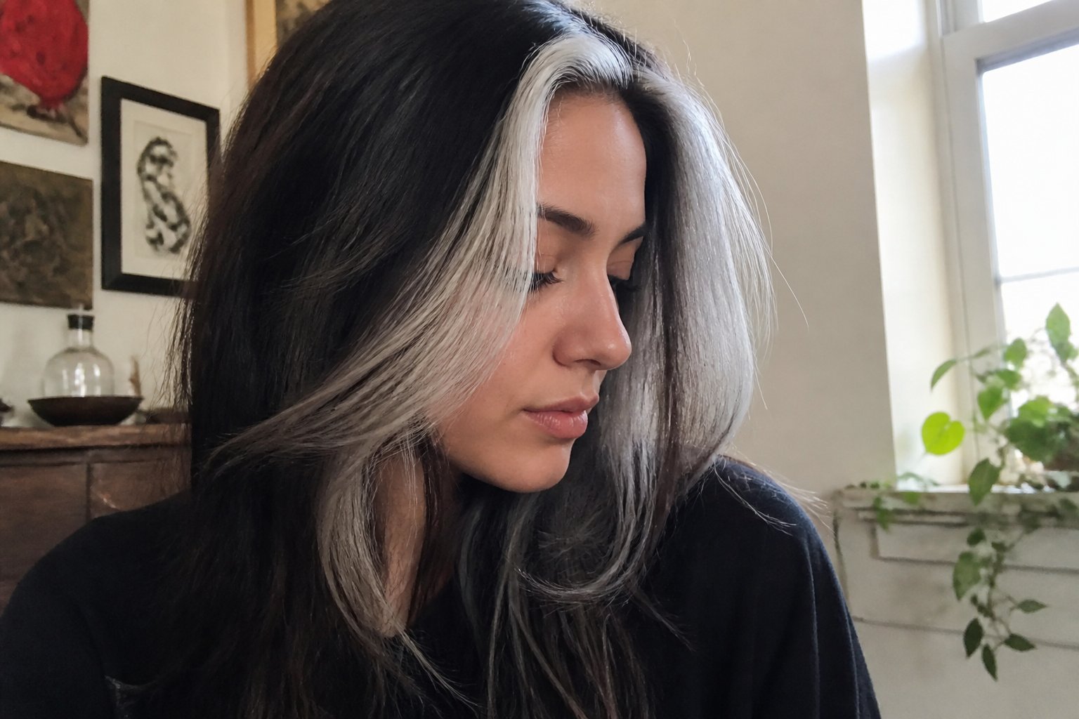 A person with a silver-gray skunk stripe hairstyle is shown in soft natural daylight, sitting casually indoors with everyday objects around them.