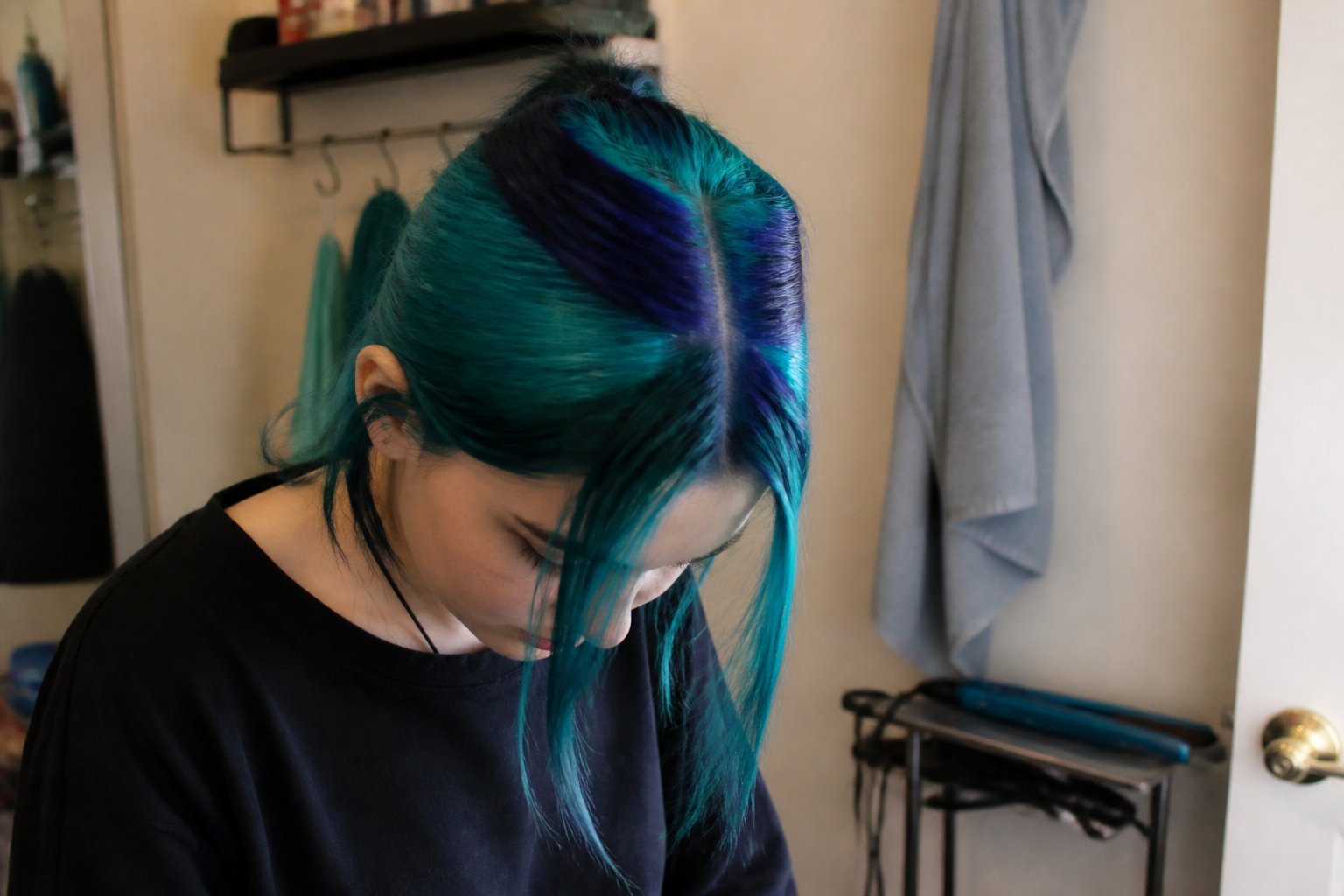 A person with teal and navy double skunk stripe hair in a casual indoor setting with hair styling tools nearby.