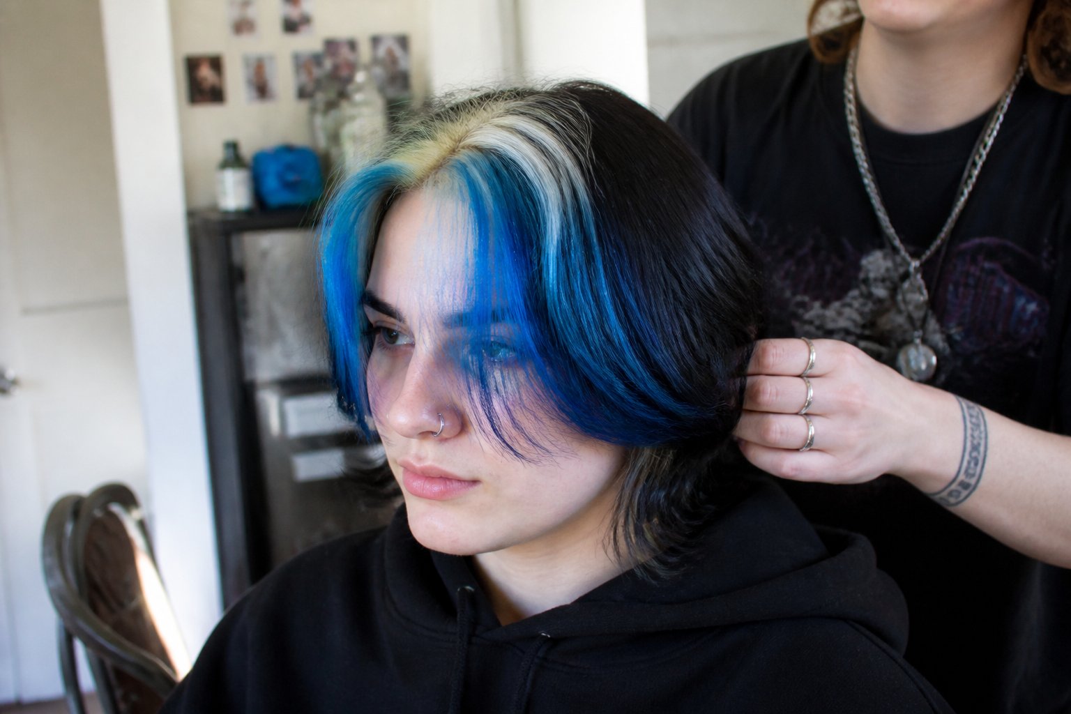 A person with electric blue skunk stripe hair is adjusting their hair with styling tools on a table nearby.