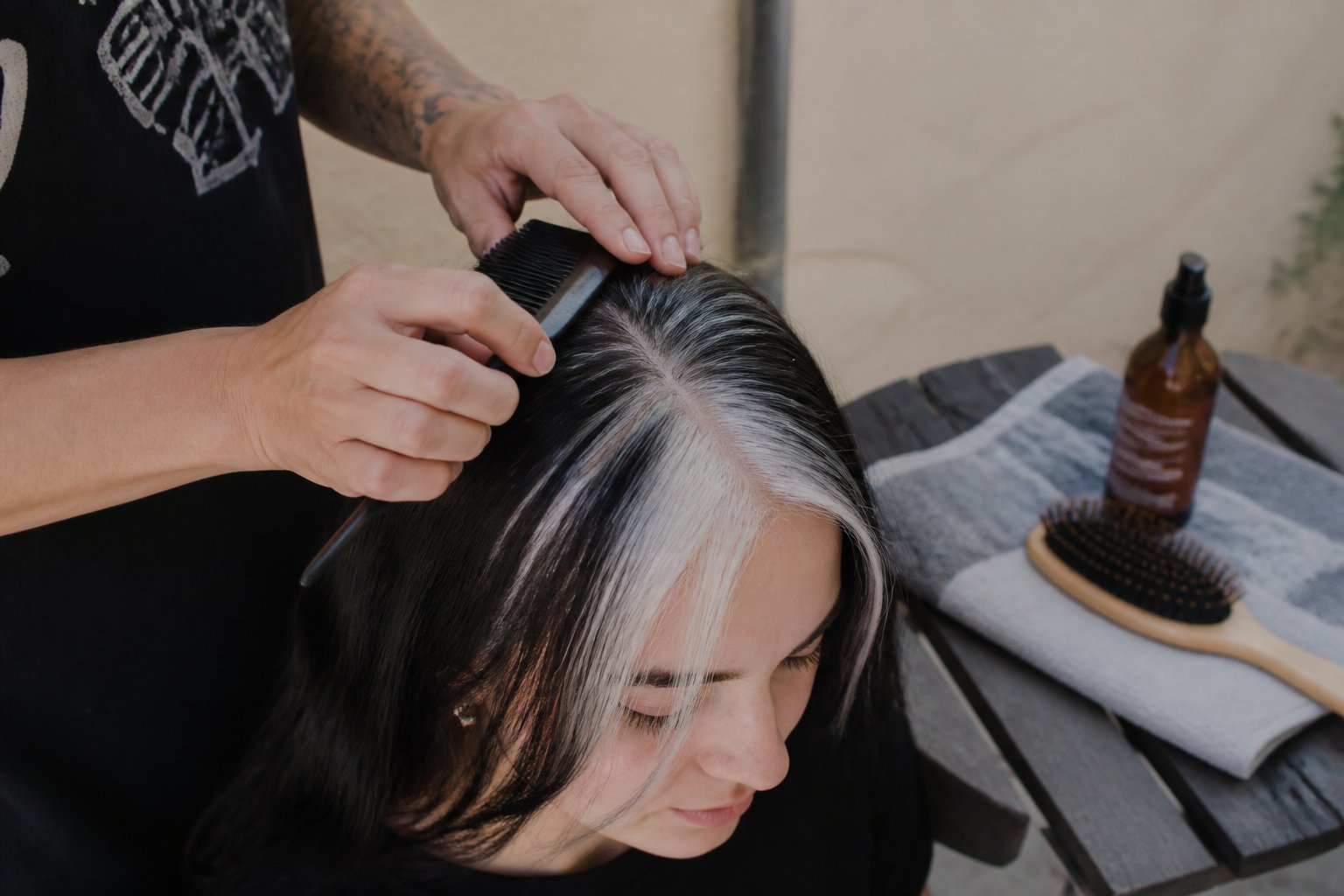 Hands caring for hair with black and white skunk stripe color, surrounded by hair care tools on a light surface.