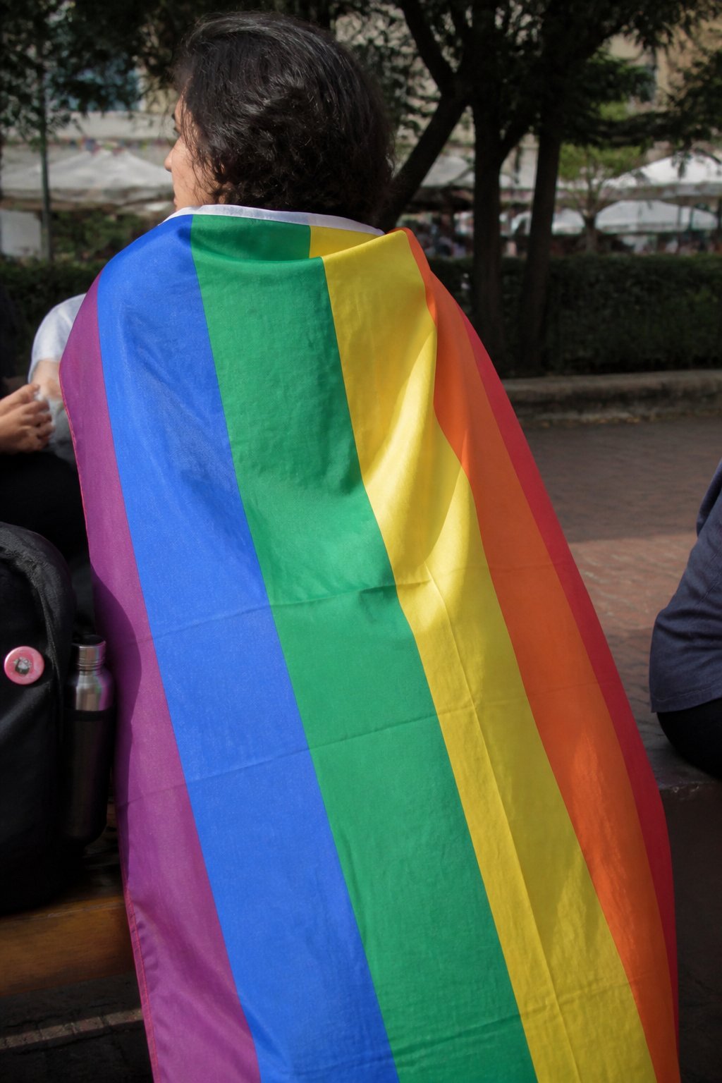Person wearing a six-color Pride flag cape outdoors in soft daylight, casually posed with everyday objects nearby.