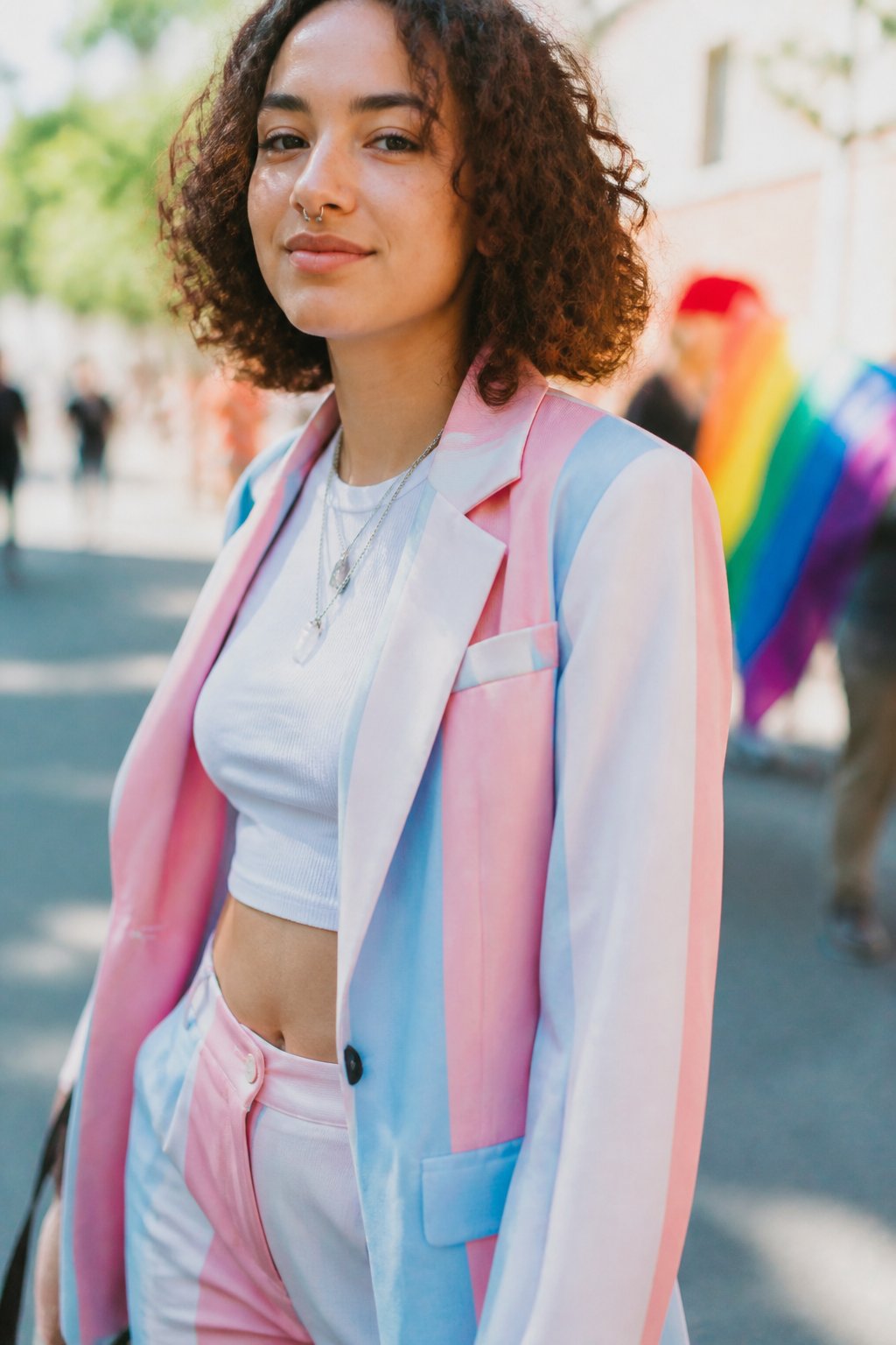 A person wearing a pastel trans flag blazer in pink, blue, and white stands casually outdoors during the day, with natural light and soft shadows around them.