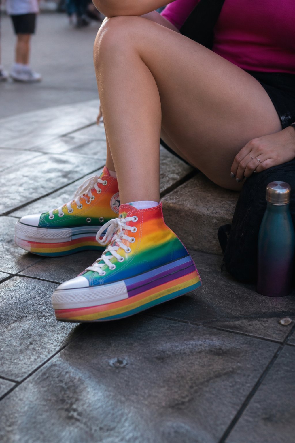 Close-up of rainbow platform sneakers worn outdoors on a sidewalk during a Pride Day outing, with a hand and personal items nearby.