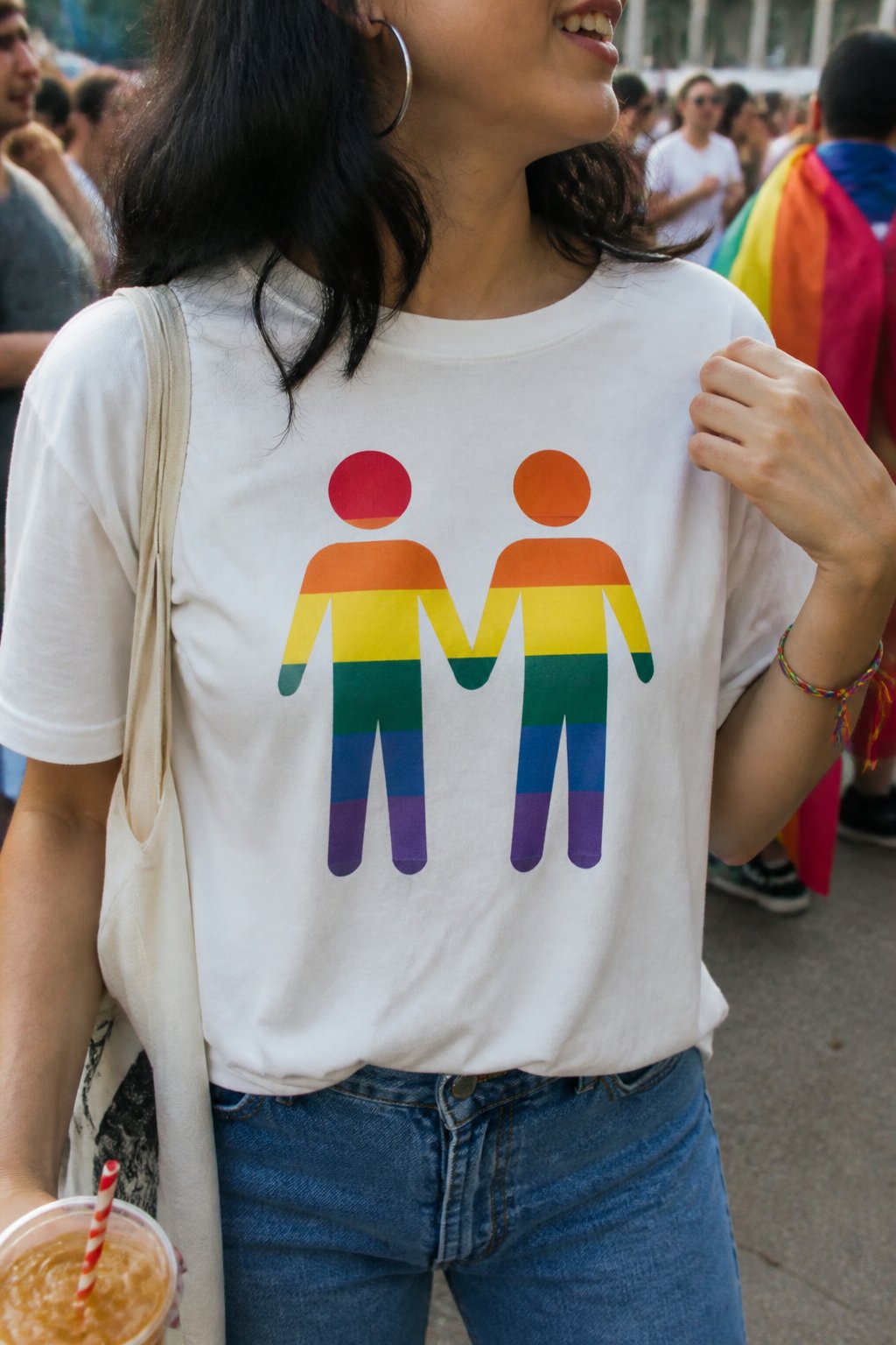 A person wearing a casual graphic tee with an inclusive slogan, standing in natural daylight with soft shadows, surrounded by everyday objects and hands adjusting the shirt.