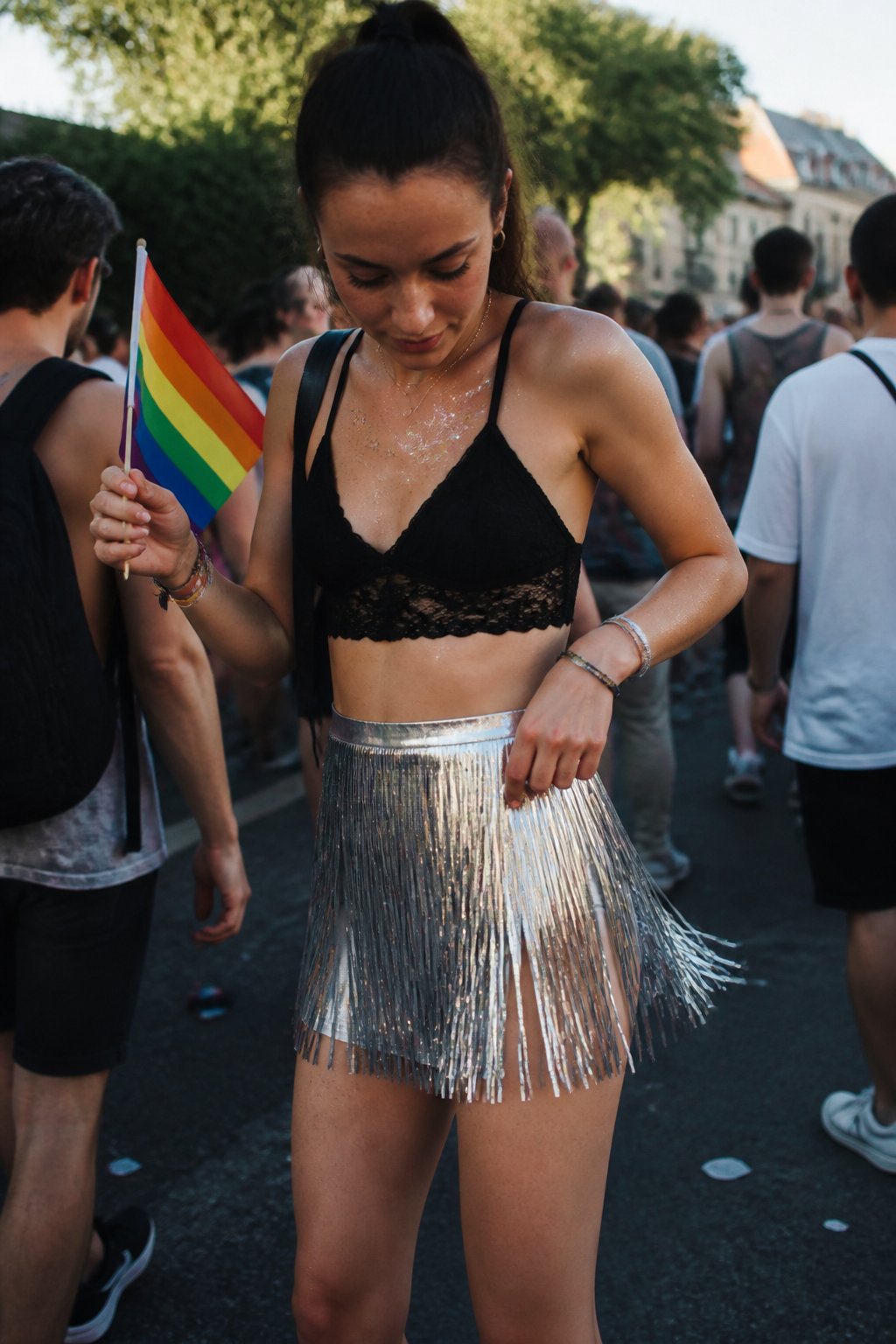A person wearing a metallic fringe skirt at a Pride Day event, with hands holding a small pride flag and casual festival accessories nearby.