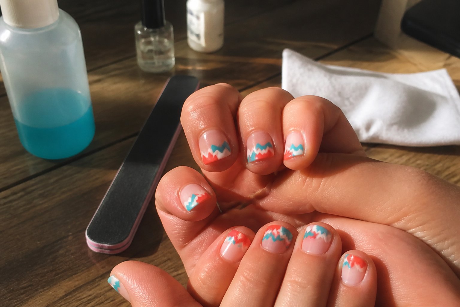 Close-up of hands with aqua and coral chevron wave tip nails, surrounded by nail care tools on a light surface.