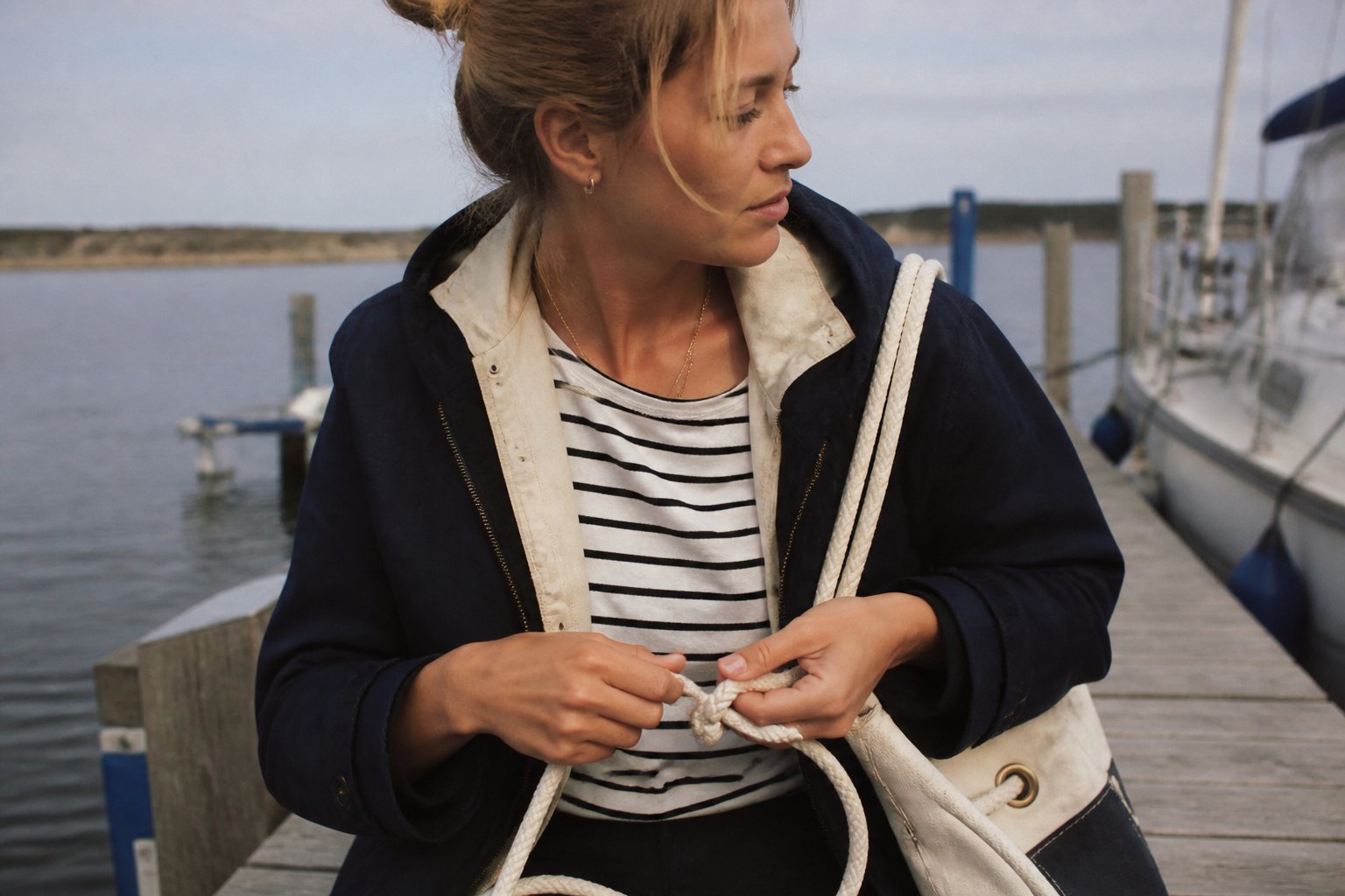 A woman in a nautical outfit stands near a wooden dock in soft daylight, holding a rope with a marina in the background.