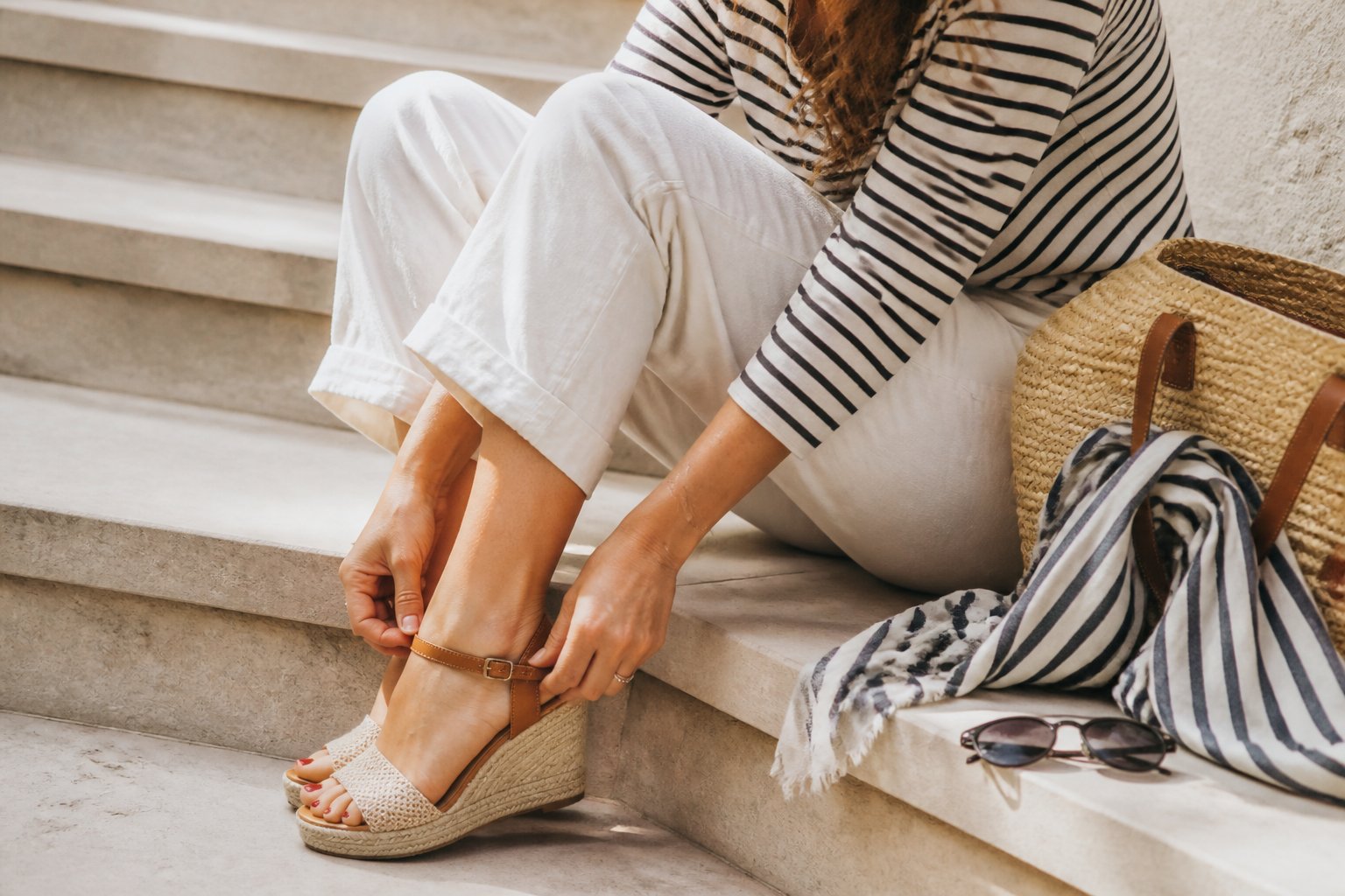 A woman wearing espadrille wedge sandals and a nautical outfit, with her hands adjusting the sandals. Natural daylight softly illuminates the scene, showing casual accessories like a striped scarf and sunglasses on a wooden surface.