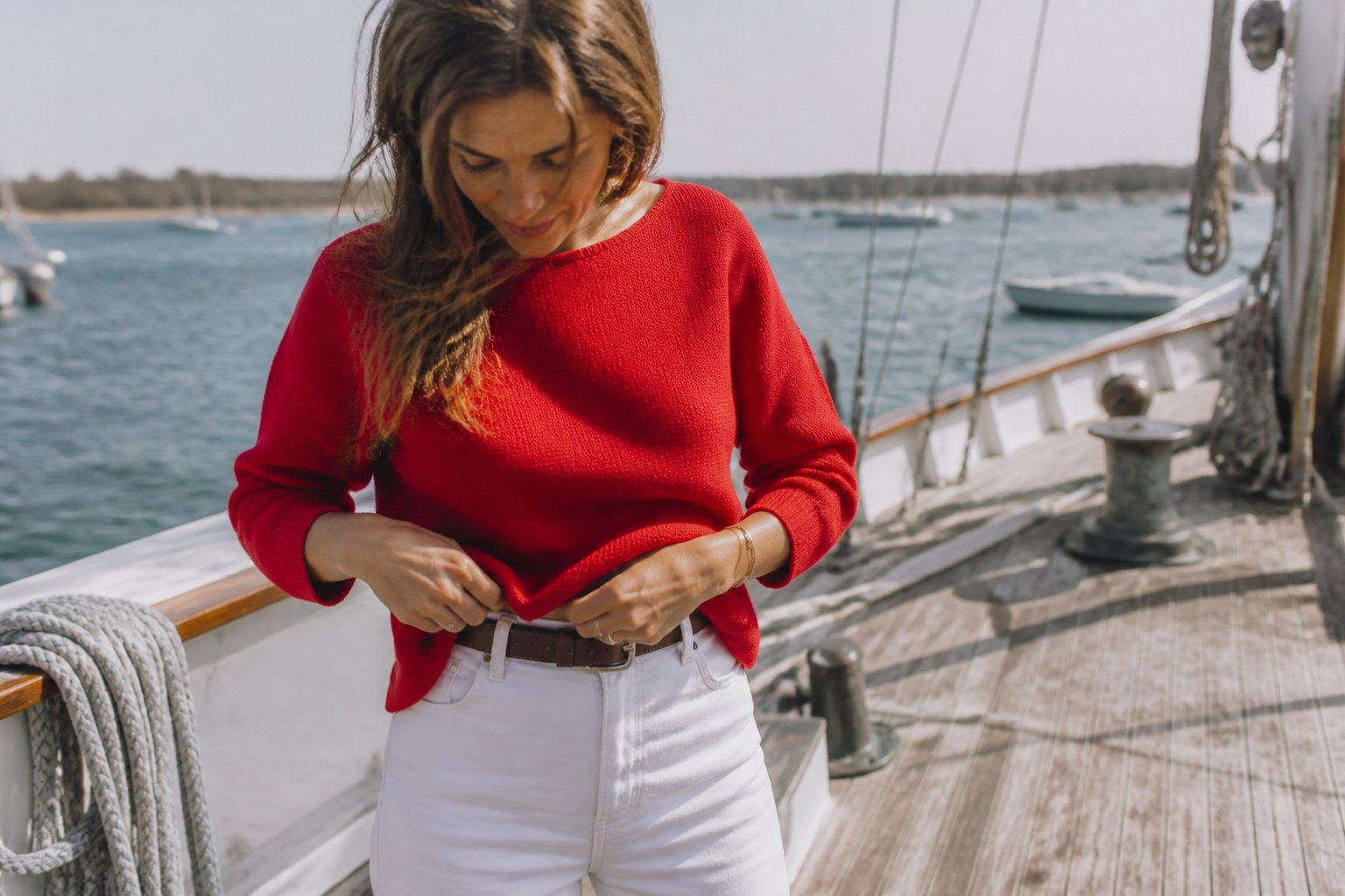 A woman wearing a red cashmere boatneck sweater stands near a wooden boat railing in soft daylight, holding a rope with a casual, relaxed posture.
