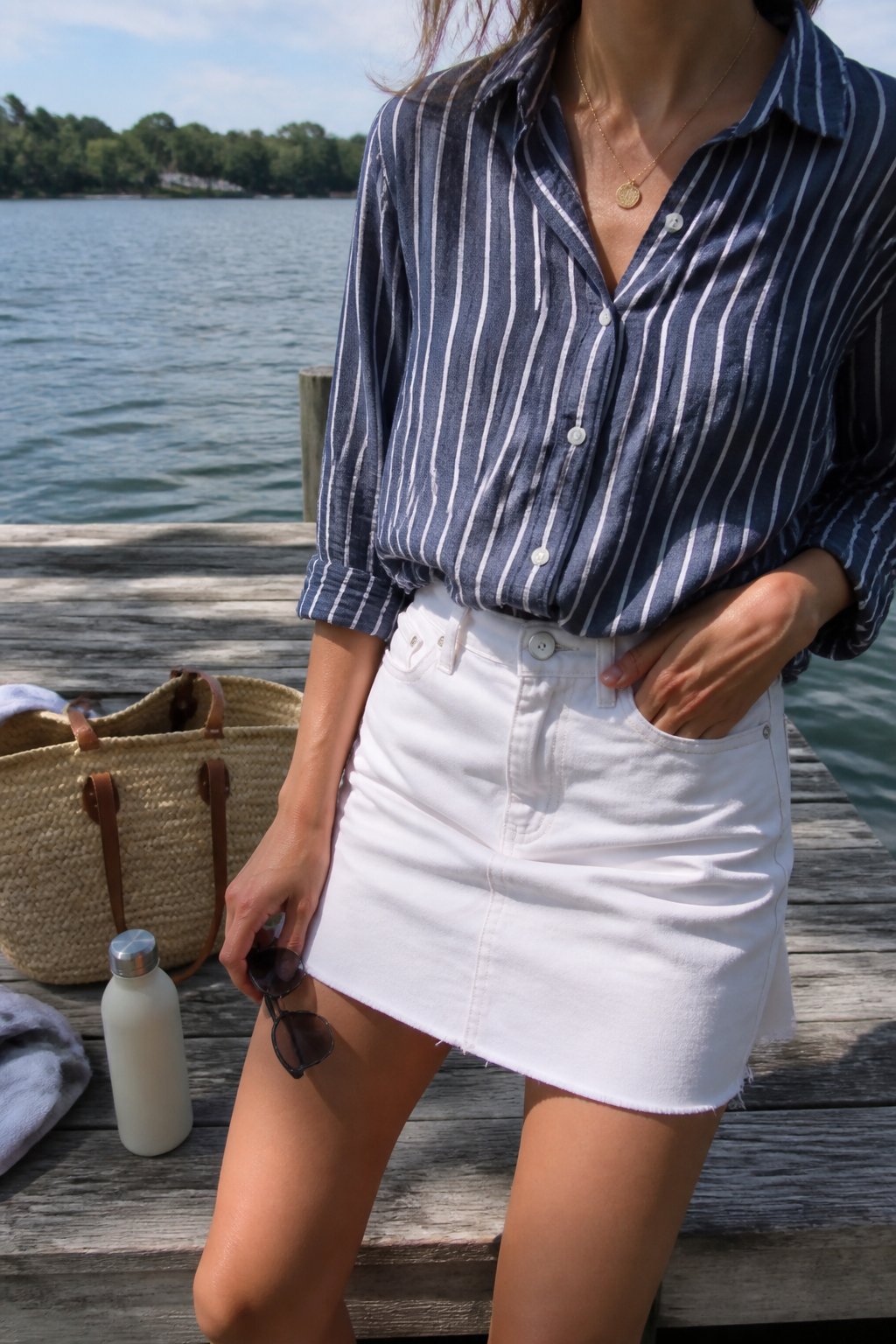 A person wearing a white denim mini skirt and navy striped linen button-down shirt standing near a wooden dock by a lake, with a woven tote bag and a water bottle nearby.