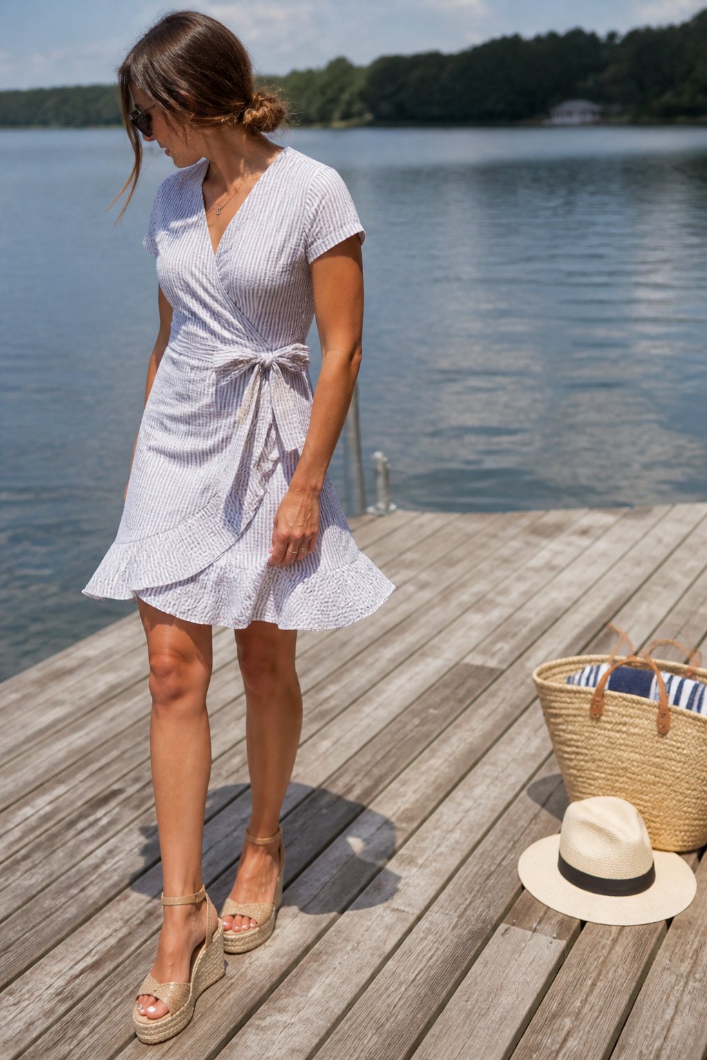 A woman wearing a seersucker wrap dress and espadrille wedges stands on a wooden dock by a lake under soft daylight.