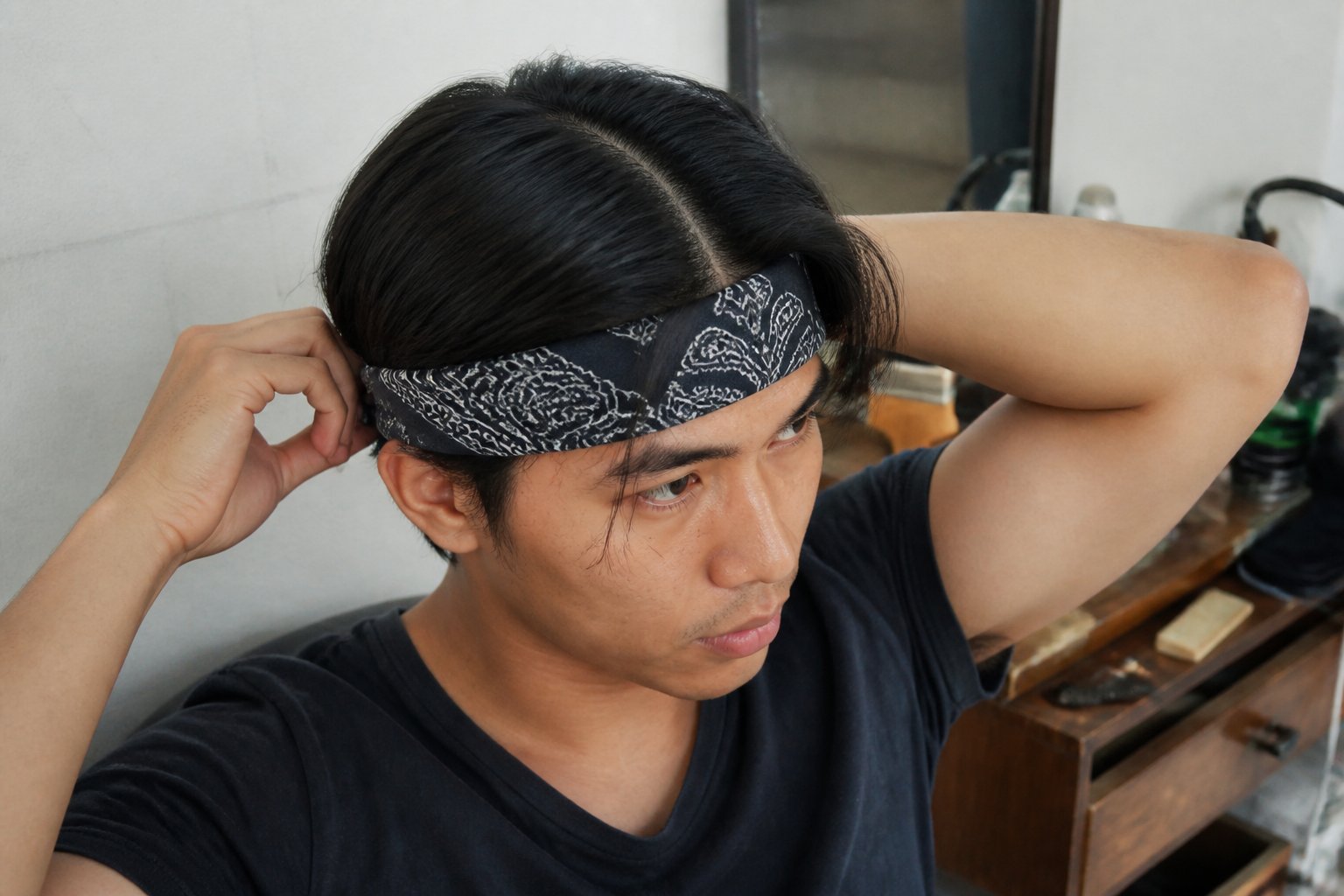 Person with a sleek middle-part hairstyle wearing a bandana as a forehead band in natural daylight, with hands and everyday objects nearby.