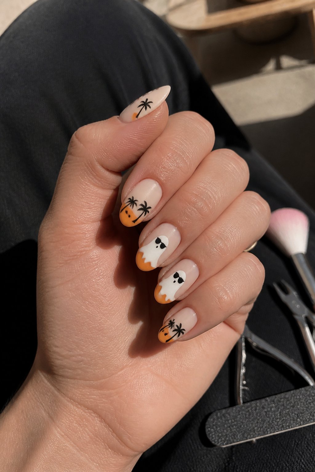 Close-up of hands with almond-shaped nails painted with ghost designs wearing sunglasses, surrounded by manicure tools on a softly lit surface.