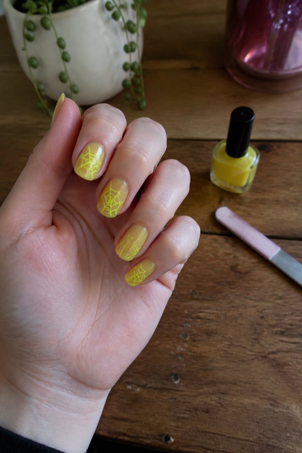 A hand with neon cobweb nail art over sheer yellow polish resting near nail care tools on a wooden surface in soft daylight.