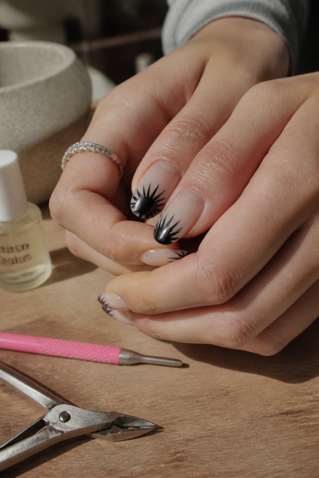 Close-up of hands with black sunburst French tip nails surrounded by manicure tools on a softly lit surface.