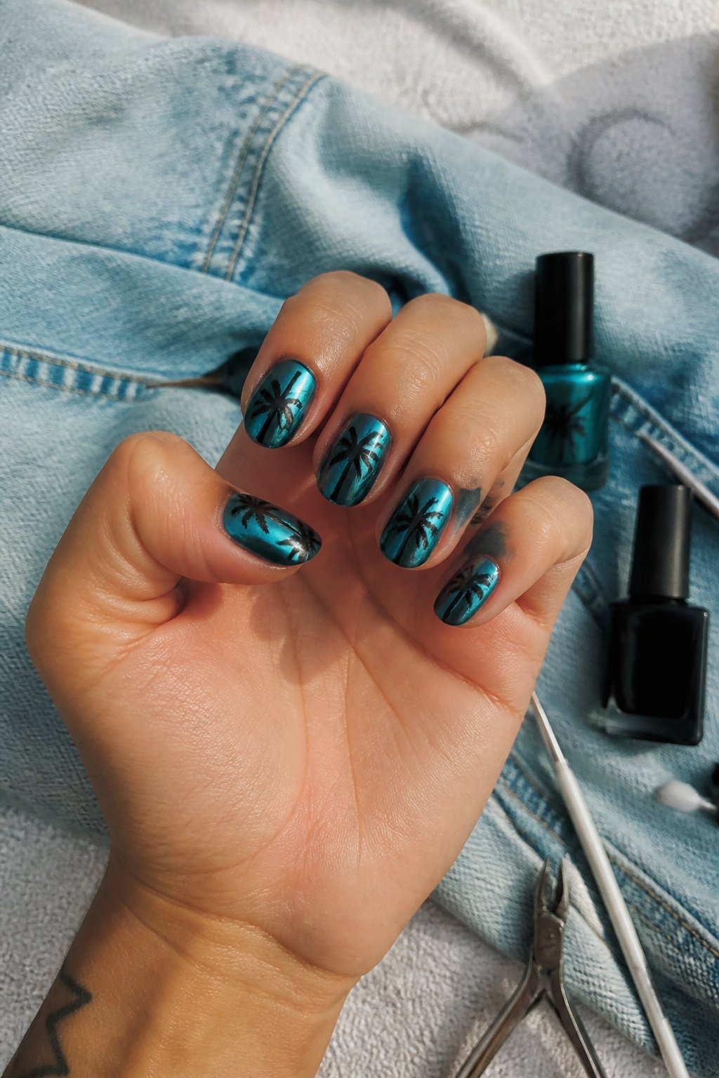 Close-up of hands with summer-themed nails near skeletal palm tree silhouettes against a teal background, with manicure tools nearby.
