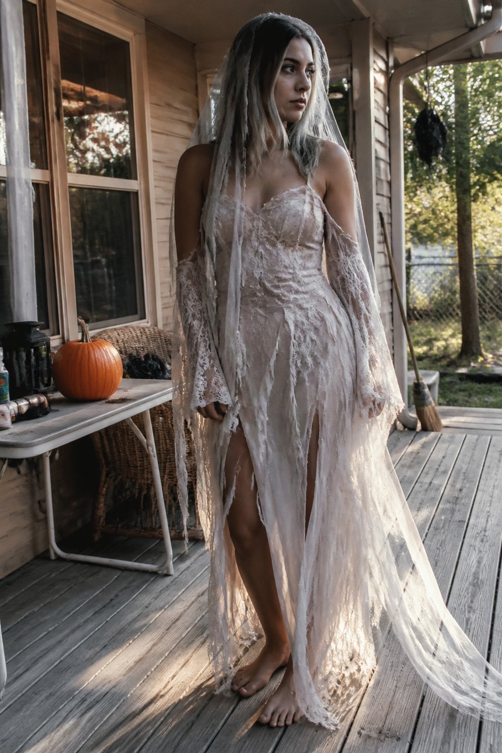 A person dressed as a ghost bride in a shredded lace dress stands outdoors with soft daylight and gentle shadows, surrounded by casual objects suggesting a relaxed setting.