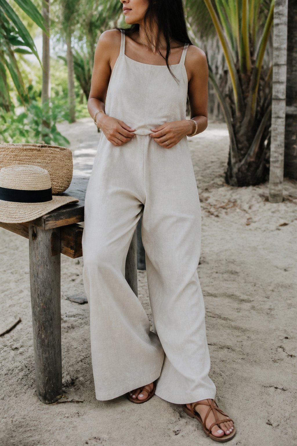 A person wearing a wide-leg linen jumpsuit and strappy sandals stands outdoors in a tropical setting with soft daylight and casual framing.