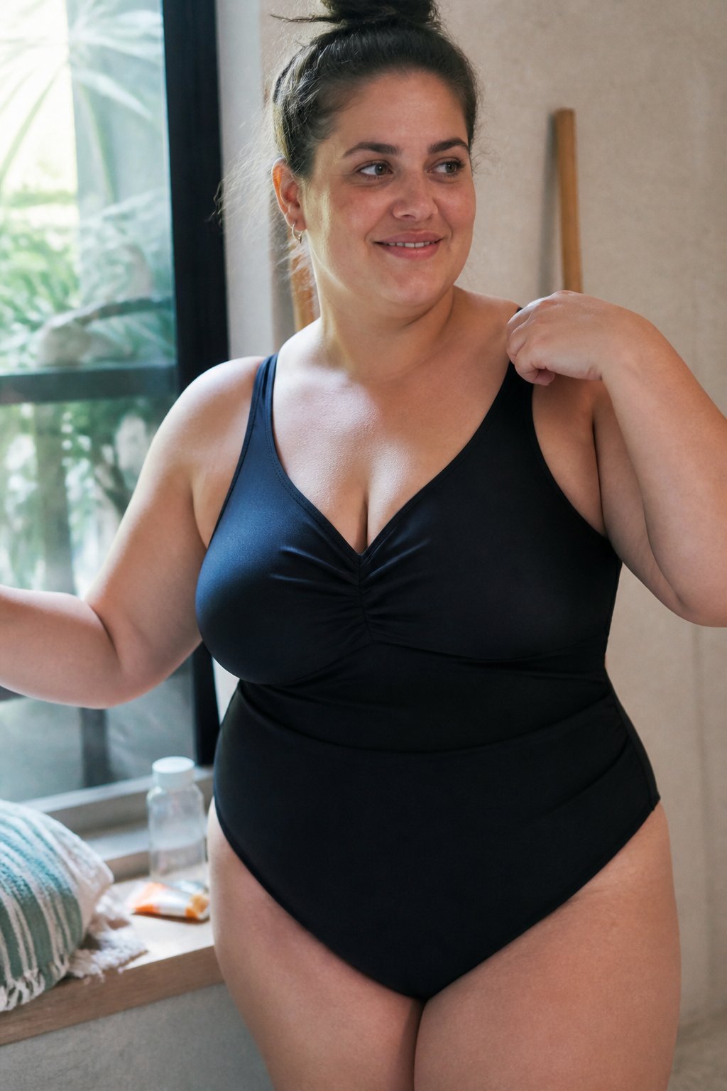 A plus size woman wearing a supportive tank bathing suit stands casually in a softly lit room with natural daylight, surrounded by everyday objects like towels and a water bottle.