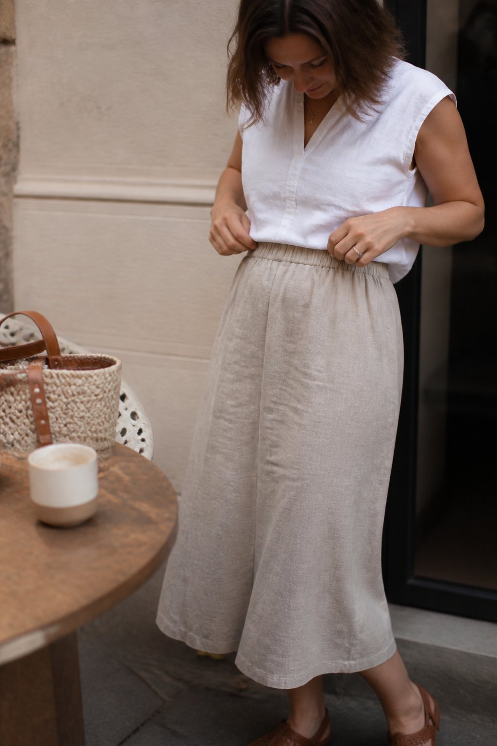 A woman wearing a high-waist A-line midi skirt with an elastic waistband stands in soft daylight with natural shadows, surrounded by everyday objects.