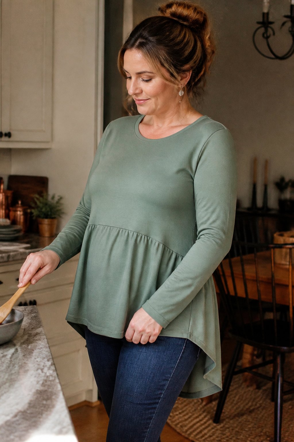 A woman wearing a peplum top with side seams and a longer back, standing casually in soft daylight with natural shadows, surrounded by everyday objects and hands in a relaxed setting.