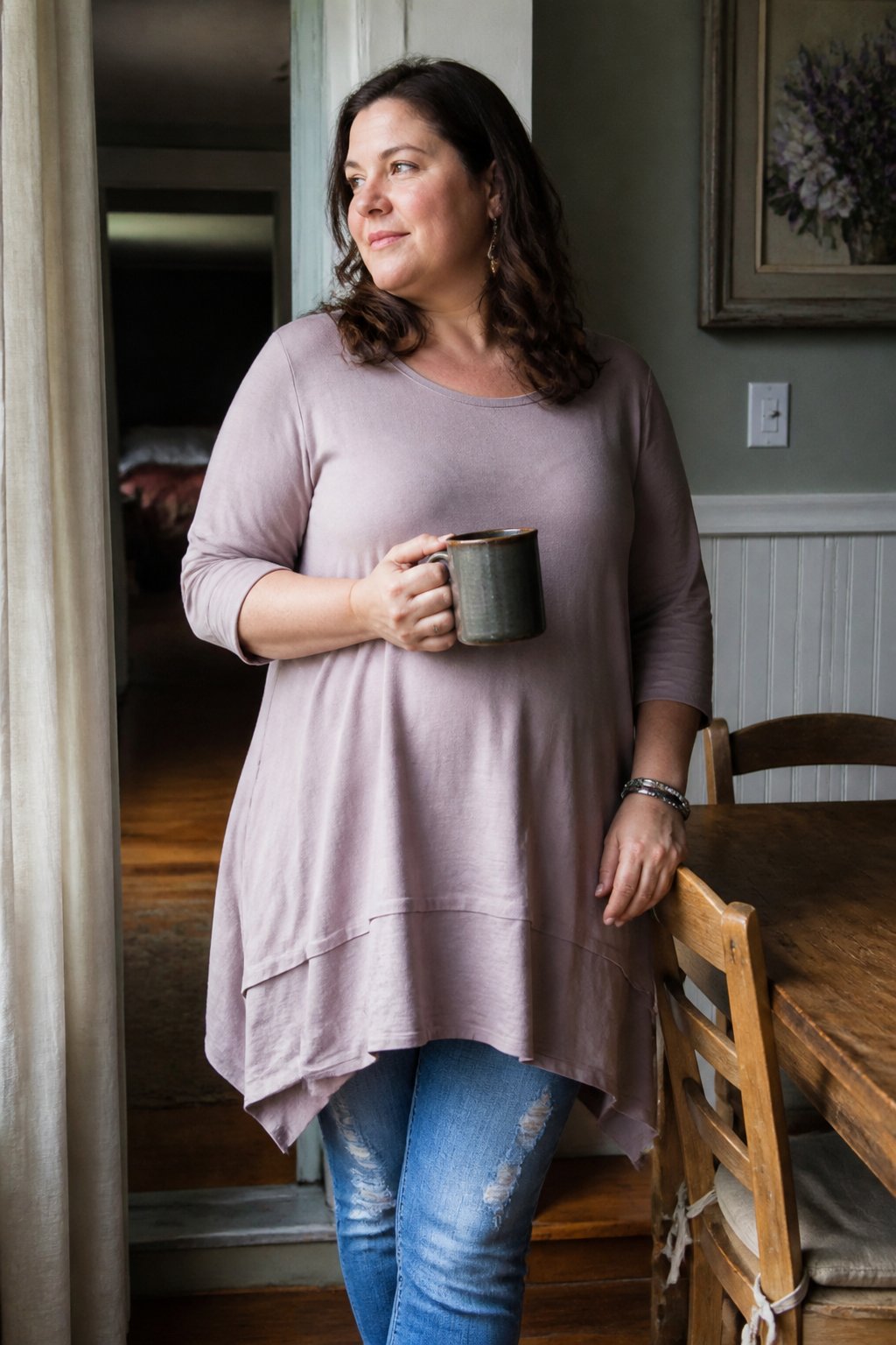 A woman wearing a tunic blouse with an asymmetric hem sits casually in a softly lit room with natural daylight, surrounded by simple everyday objects.