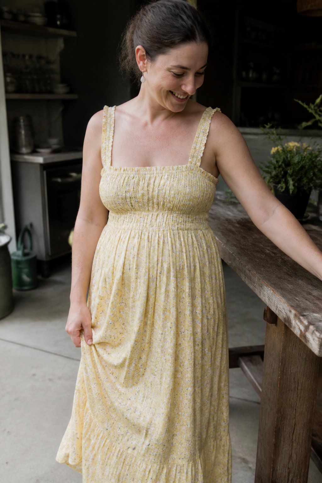 A woman wearing a shirred bodice dress with a full skirt stands in soft daylight, with natural shadows and casual framing. Hands and everyday objects are visible nearby.