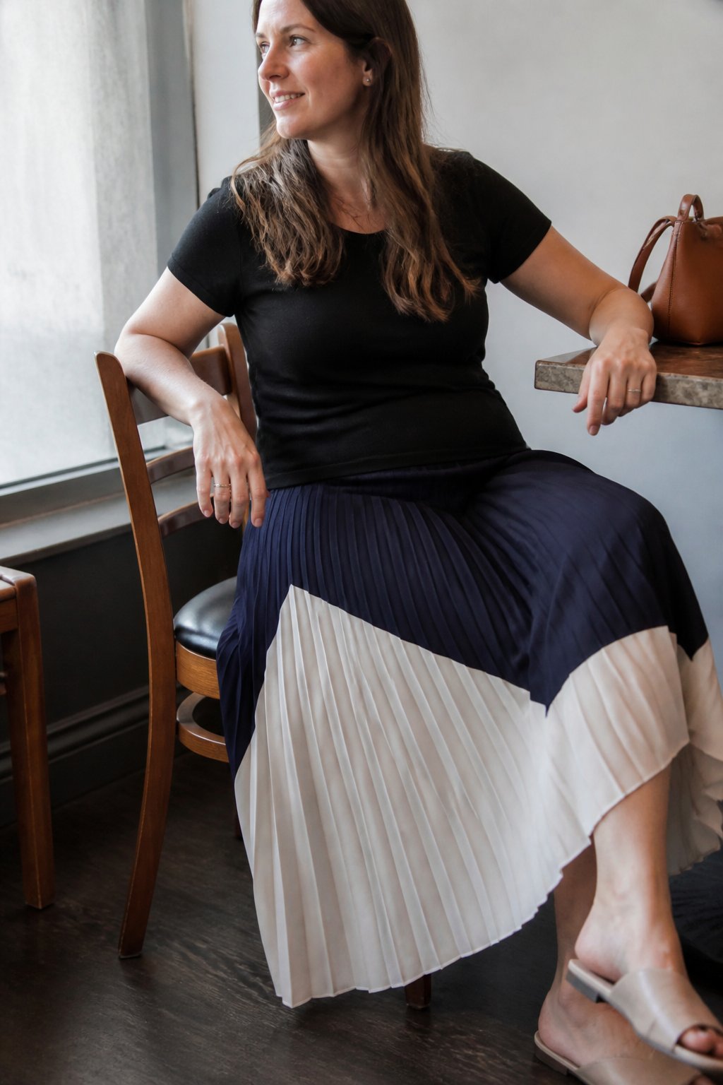 A woman wearing a pleated midi skirt with opaque panels is standing in a softly lit room with natural daylight, surrounded by everyday objects and a casual setting.