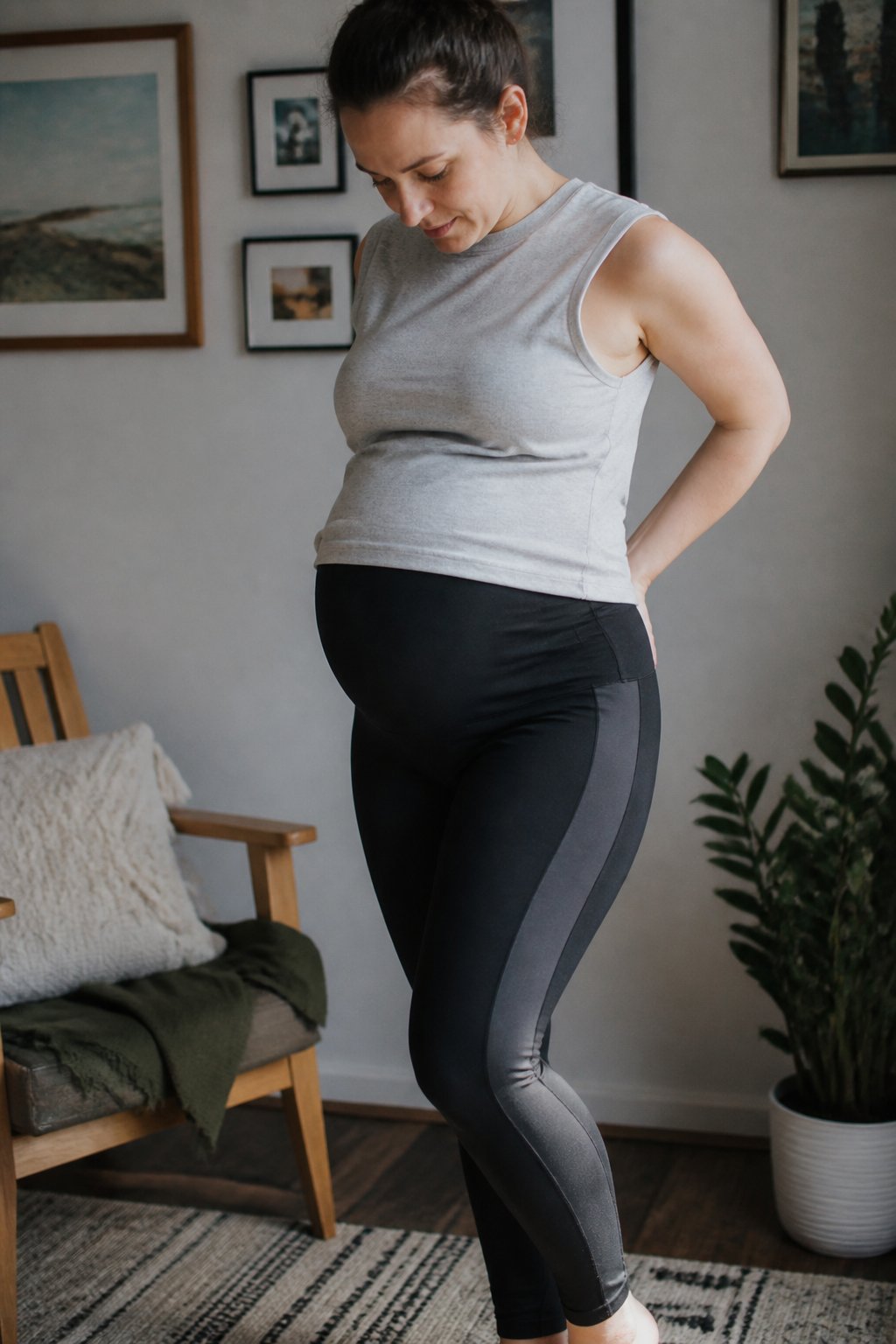 A pregnant woman wearing high-waist maternity leggings with side panels sits casually in a softly lit room with natural daylight, surrounded by simple home objects.