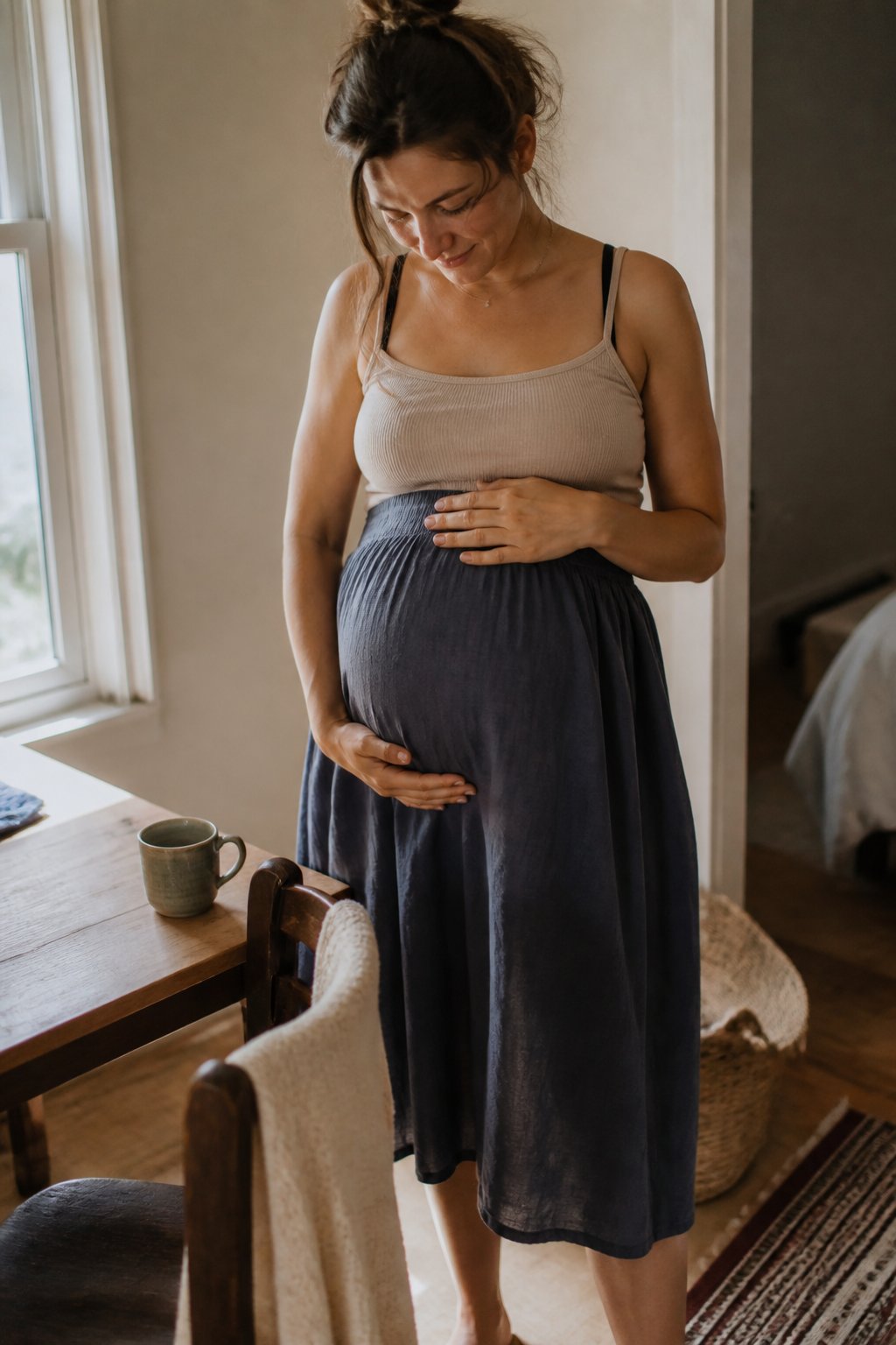 Pregnant woman wearing an elastic-waist midi skirt and maternity camisole in a softly lit room with natural daylight, surrounded by everyday home items.