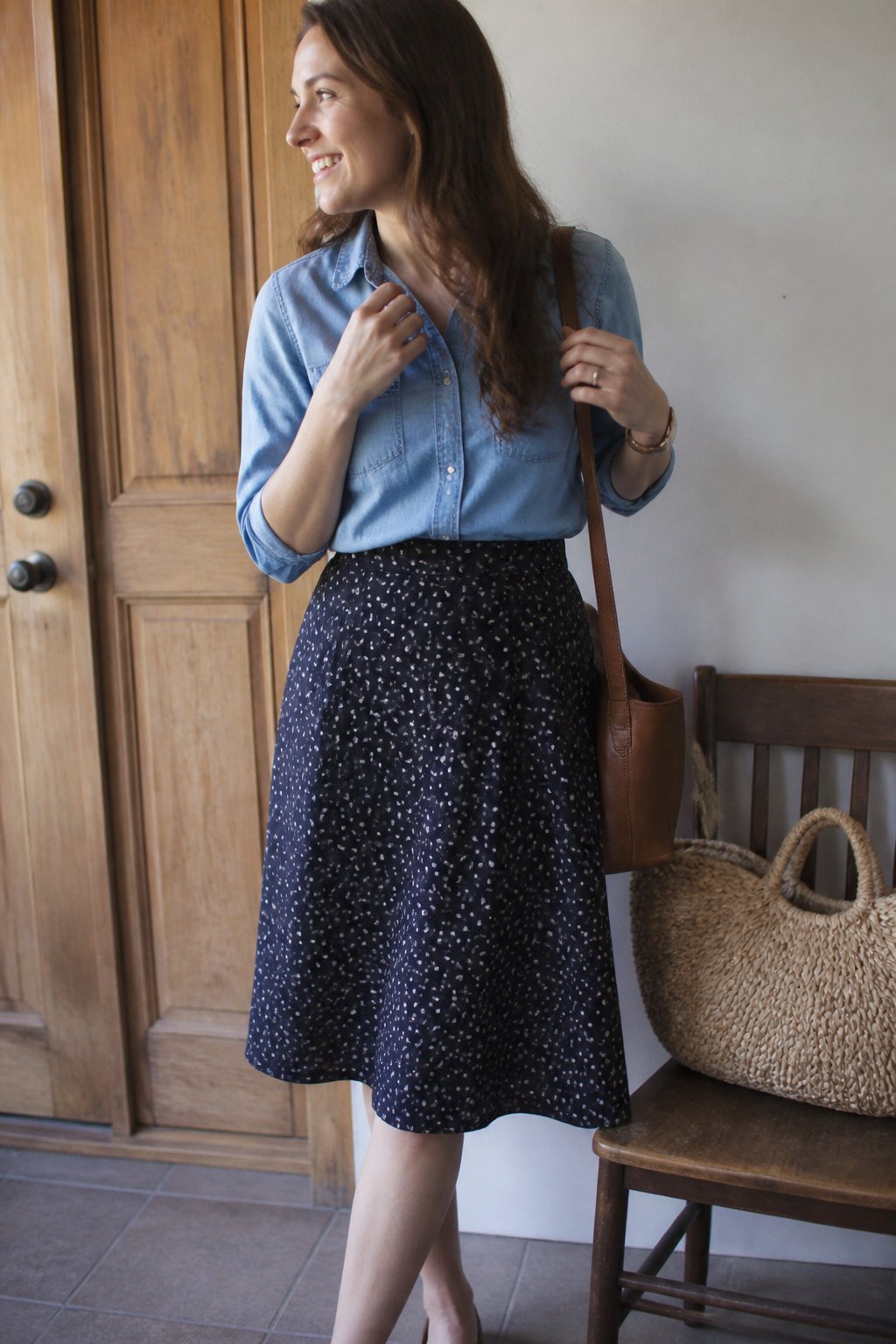 A woman wearing a knee-length A-line skirt and a tucked-in chambray shirt standing indoors with soft daylight, casual and natural surroundings.